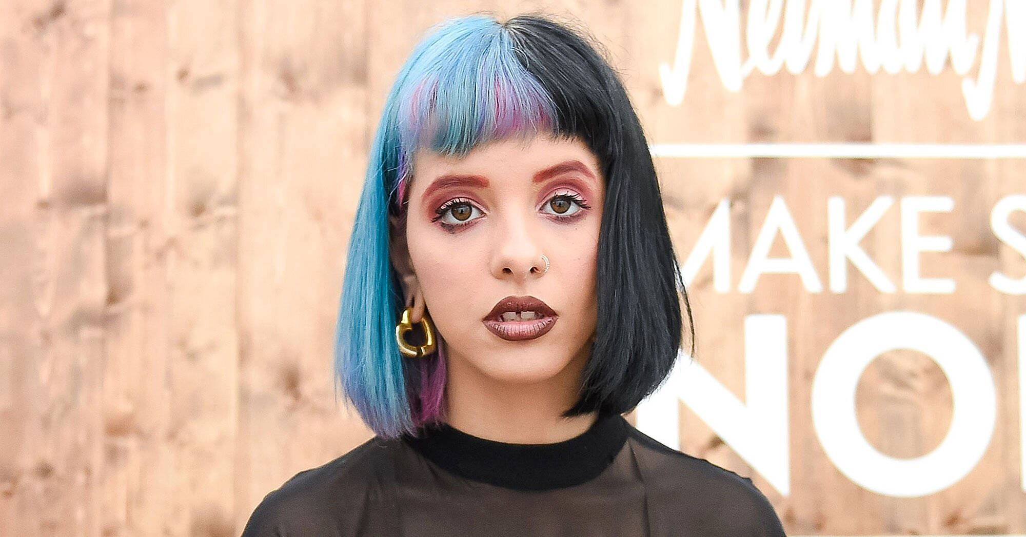 Melanie 桌布 壁紙 — 一位年輕女性，擁有搶眼的藍黑髮色和引人注目的妝容，在帶有白色文字的木質背景前擺姿勢。