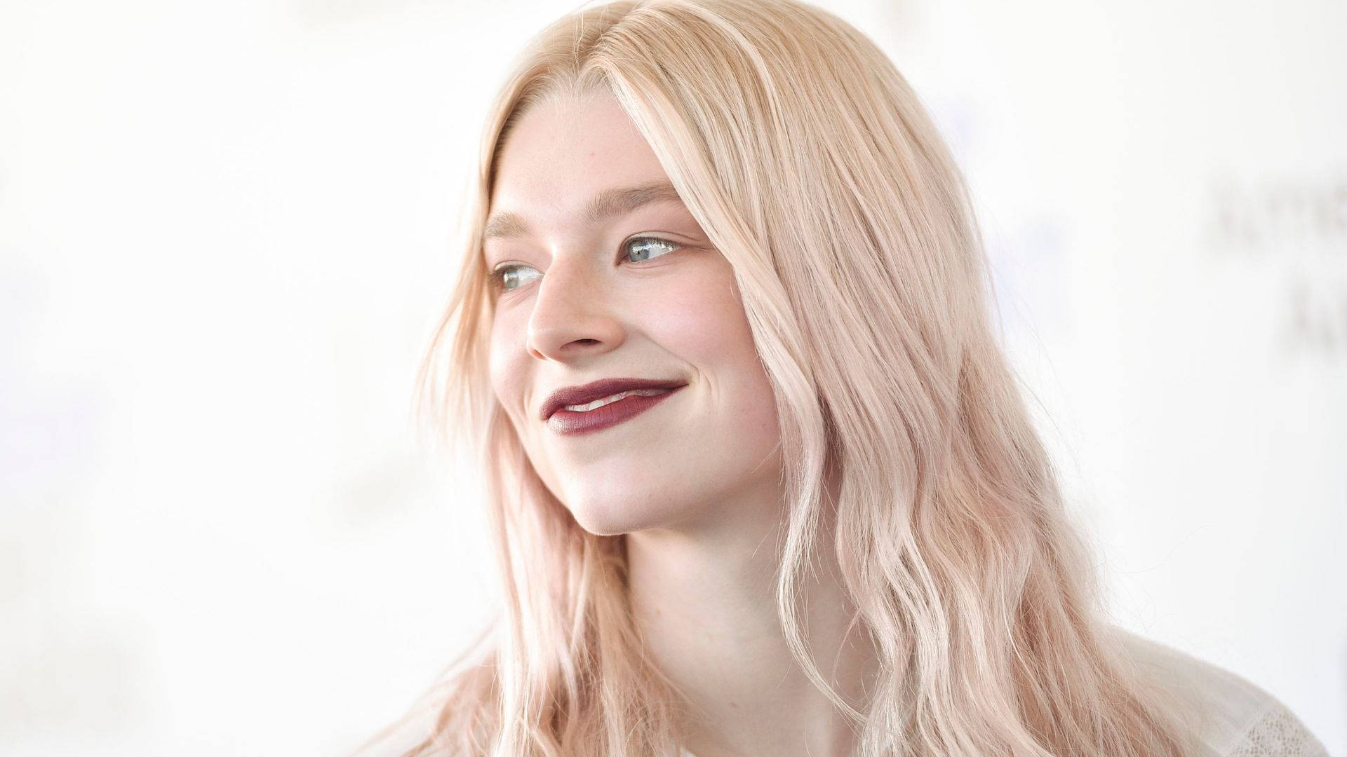 Hunter Schafer 桌布 壁紙 — 一位年輕女性，擁有柔軟的金色頭髮和溫柔的微笑，在明亮的白色背景上若有所思地望向一旁。