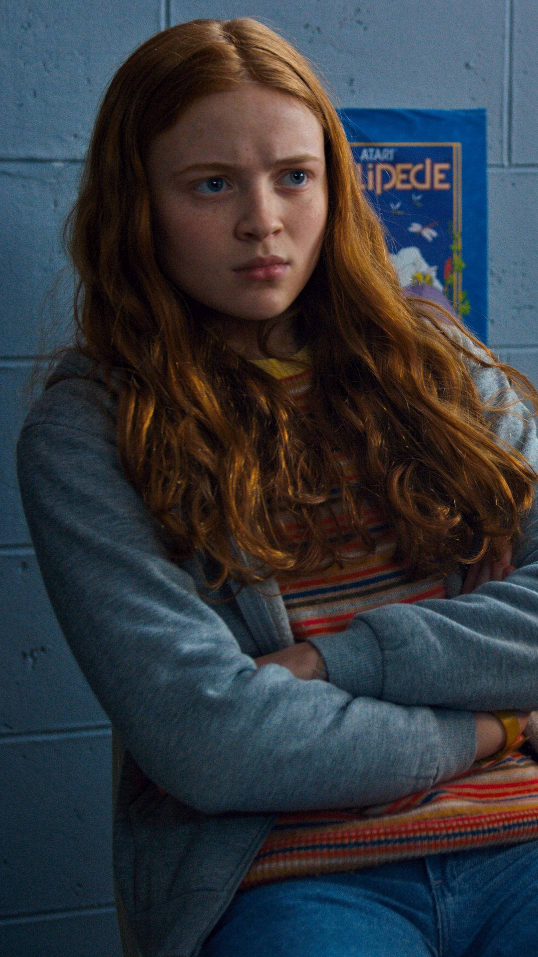 Stranger Things iPhone 桌布 壁紙 — 一位年輕女性，擁有火紅的頭髮和懷疑的表情，雙臂交叉站在藍色煤渣磚牆前。