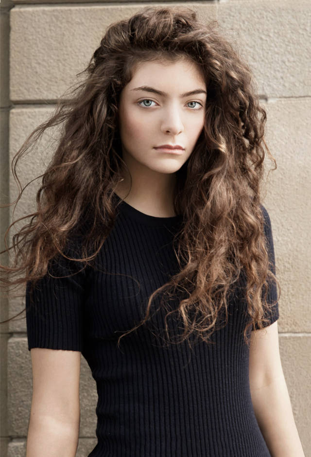 Lorde 桌布 壁紙 — 一位年輕女性，擁有豐盈的棕色捲髮和引人注目的藍色眼睛，在紋理石牆前直視鏡頭。