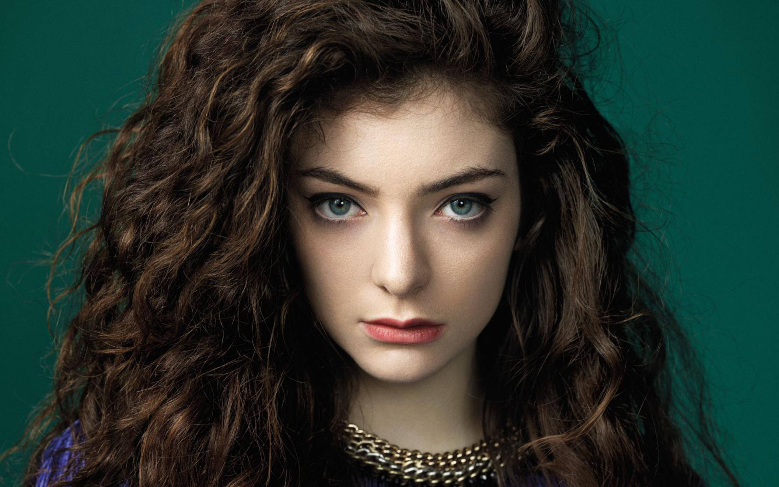 Lorde 桌布 壁紙 — 一位年輕女性，擁有豐盈的棕色捲髮和引人注目的藍色眼睛，在純色藍綠色背景下凝視著。