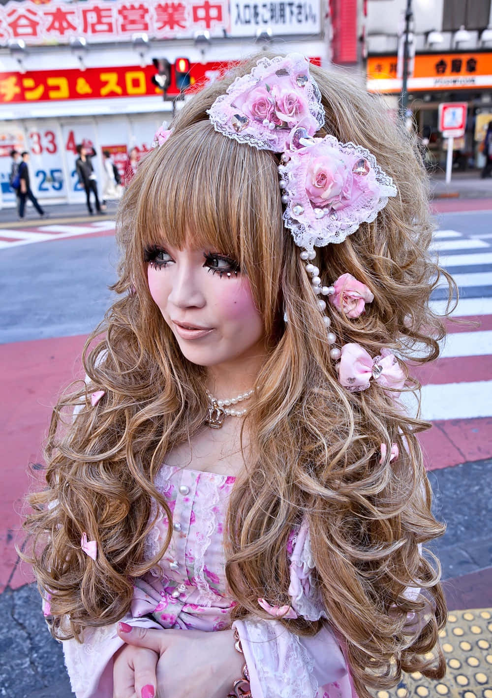 Gyaru 桌布 壁紙 — 一位年輕女性，擁有豐盈的金色捲髮和粉紅色花卉髮飾，站在繁忙的東京街頭，穿著相配的粉紅色服裝。