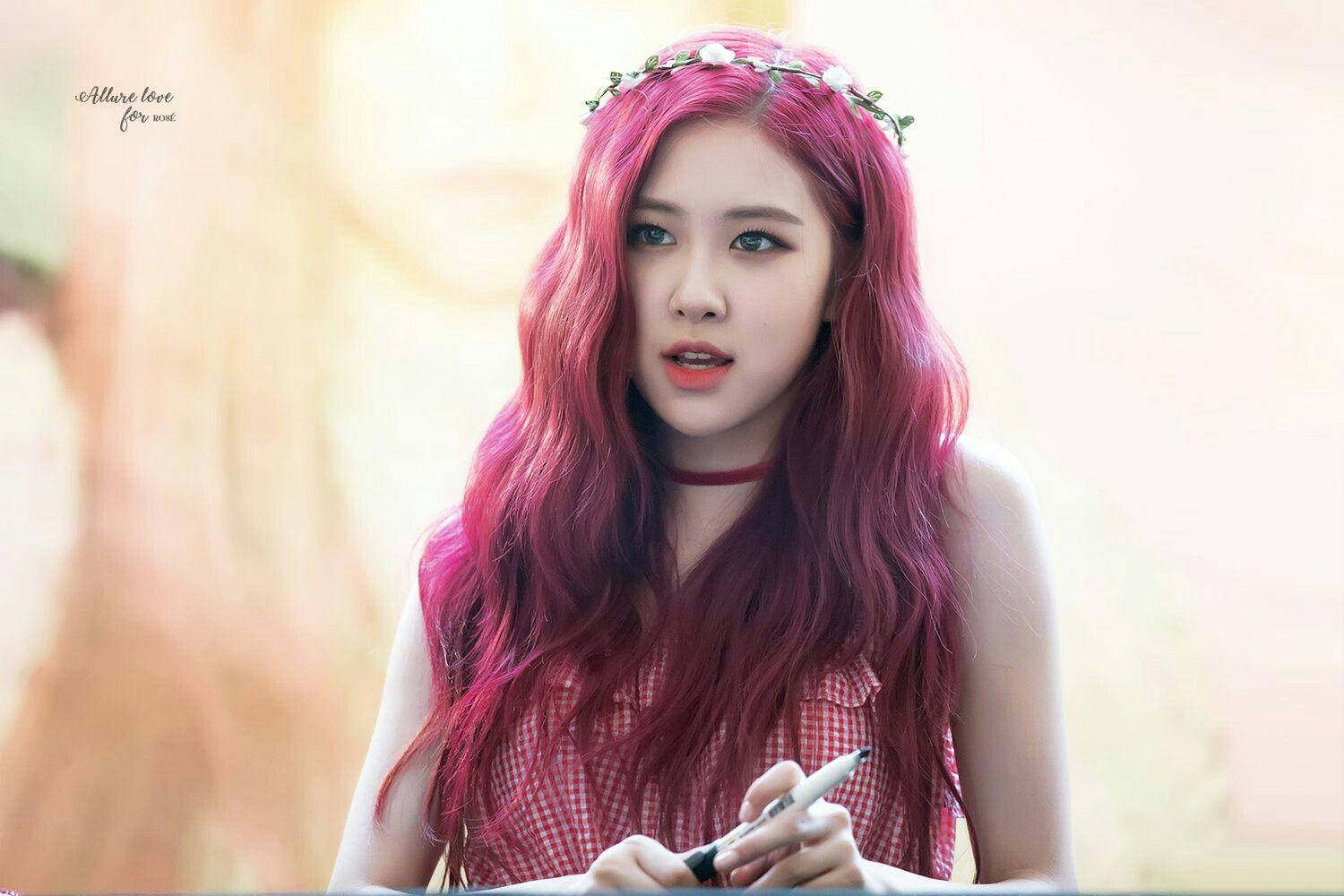 blackpink rose 桌布 壁紙 — 一位年輕女性，擁有鮮豔的粉紅色頭髮和花冠，握著一支筆，若有所思地向前看。