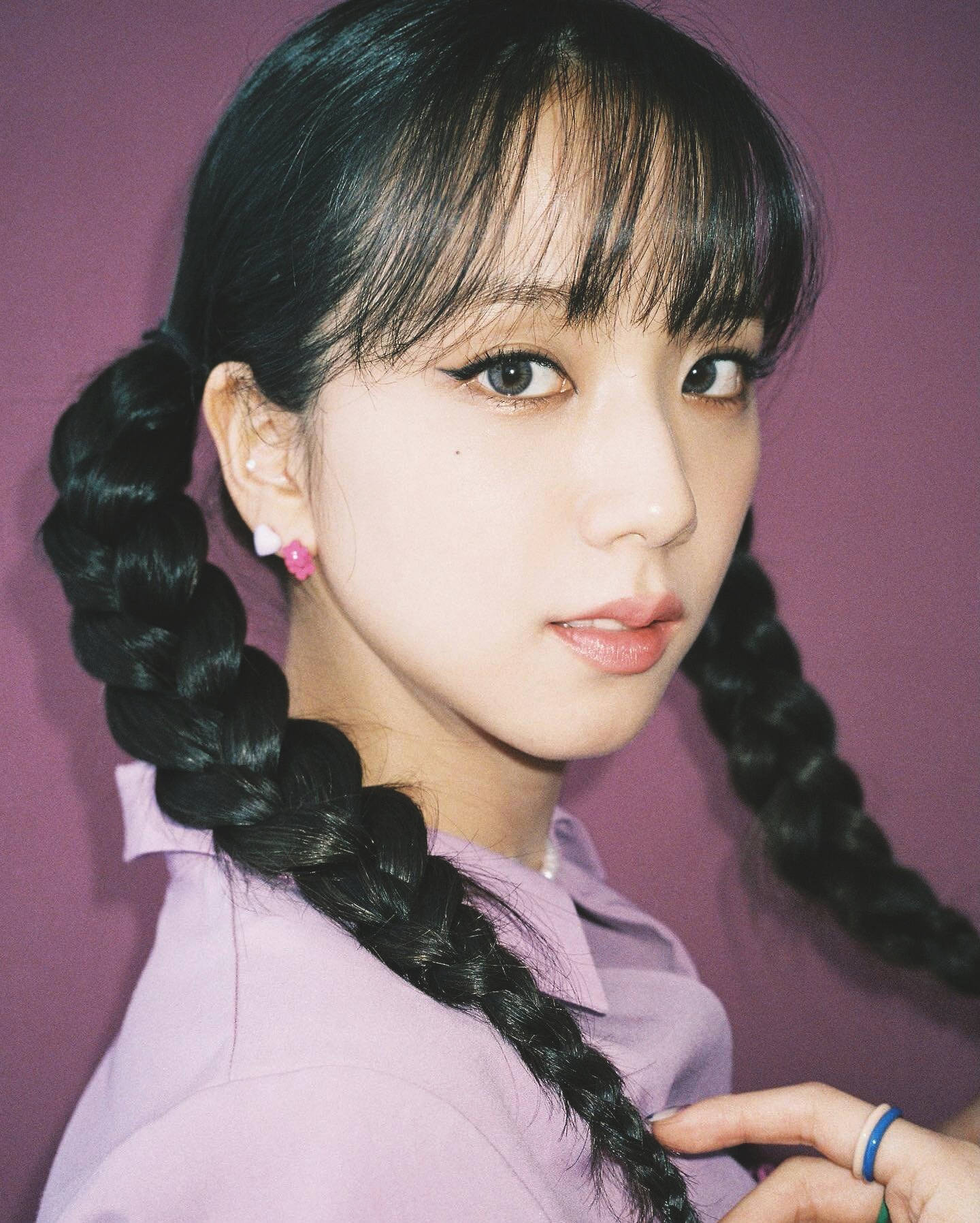 jisoo 可愛 桌布 壁紙 — 一位年輕女性，梳著粗辮子，戴著粉紅色飾品，在純紫色背景前溫柔地凝視。