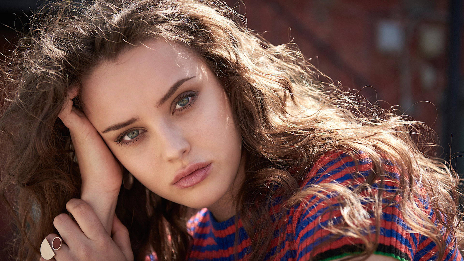 Katherine Langford 桌布 壁紙 — 一位年輕女性，棕色亂髮，在陽光普照的城市環境中，用手托著頭，專注地凝視鏡頭。