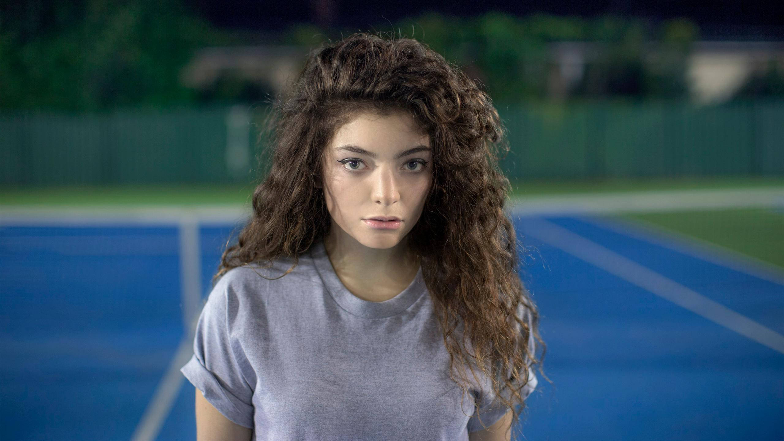 Lorde 桌布 壁紙 — 一位年輕女性，棕色捲髮，在夜間燈光下的藍色網球場上，專注地凝視著相機。
