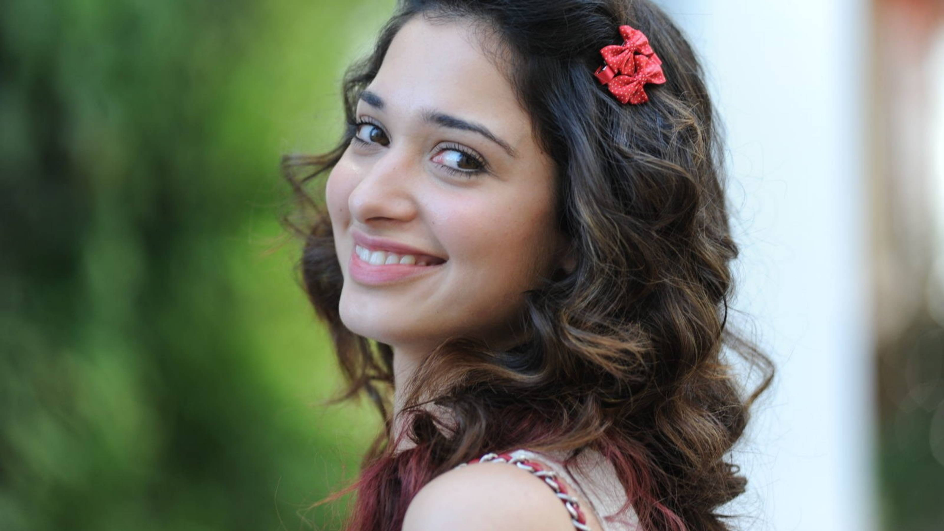 tamannaah bhatia 桌布 壁紙 — 一位年輕女性，棕色波浪長髮，戴著紅色花朵髮夾，在柔和的綠色模糊背景前對著鏡頭溫暖微笑。