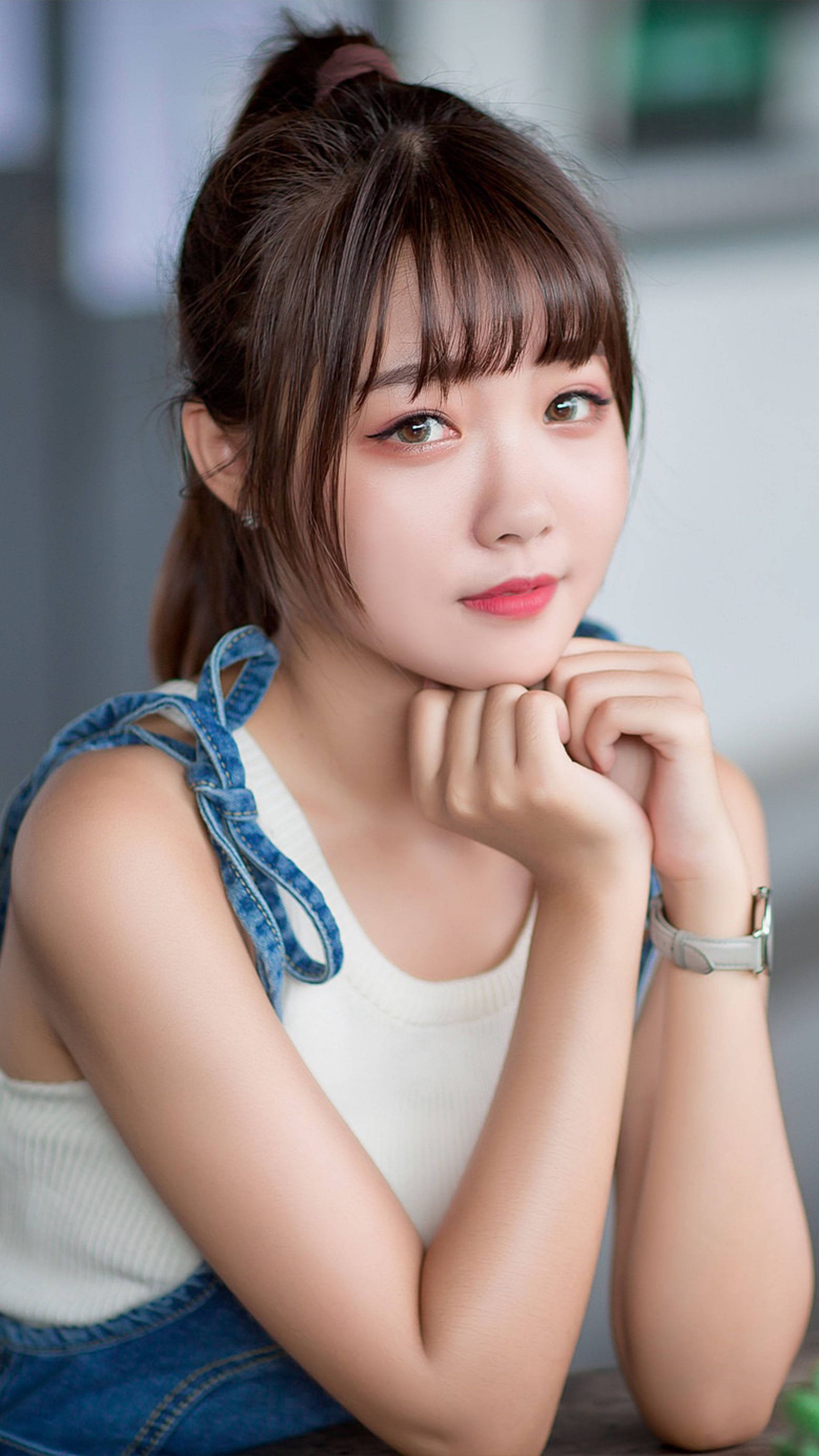 Cute Girl 桌布 壁紙 — 一位年輕女性，棕色頭髮紮成高馬尾，穿著白色上衣和牛仔吊帶褲，雙手托腮。