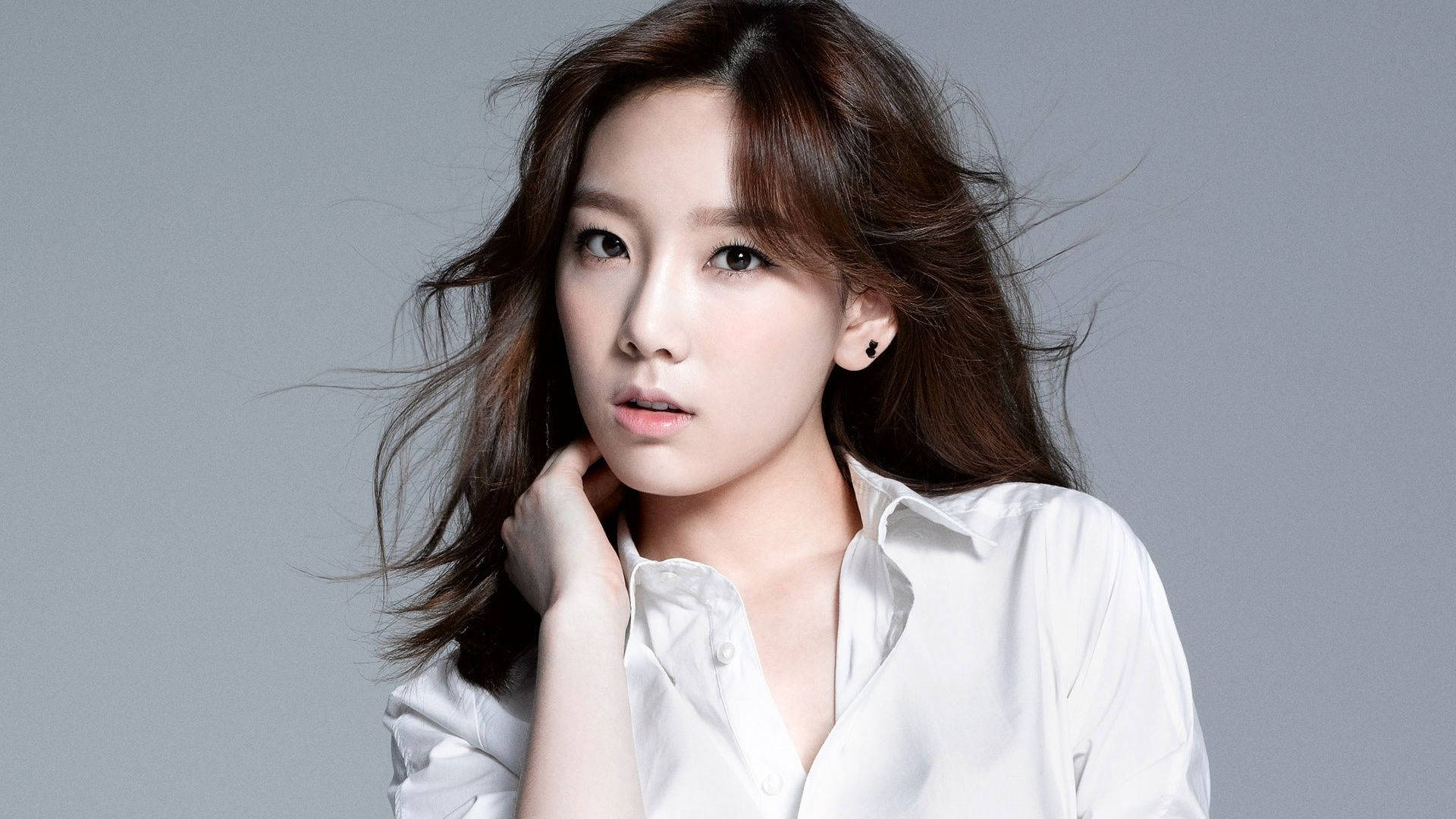 Taeyeon 桌布 壁紙 — 一位年輕女性，棕色頭髮被風吹亂，身穿白色襯衫，在純灰色背景前若有所思地凝視。