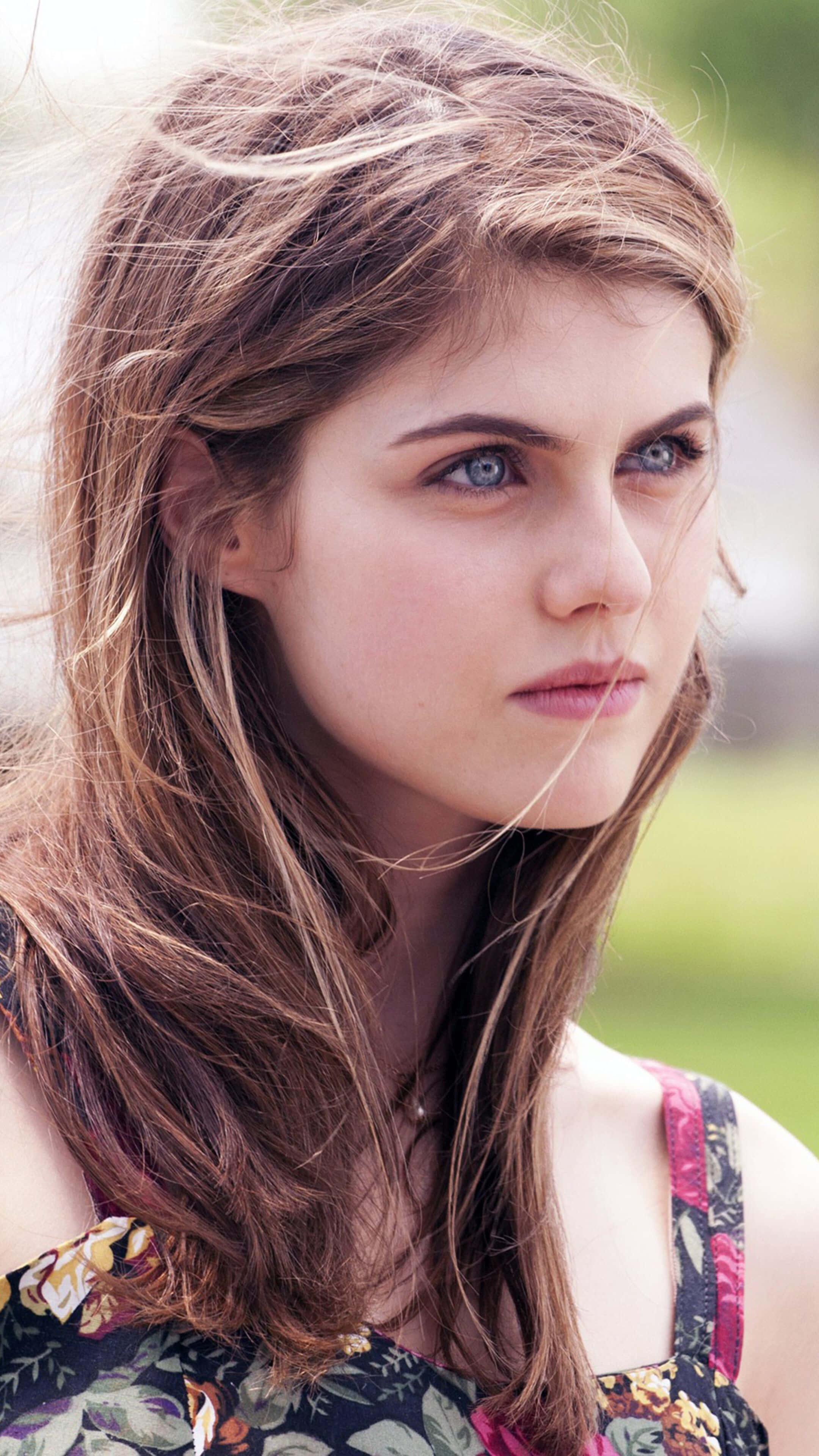 Alexandra Daddario 桌布 壁紙 — 一位年輕女性，棕髮隨風飄揚，藍眼睛銳利，在柔和的自然光下若有所思地凝視戶外。