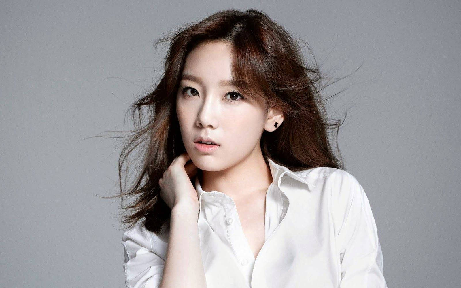 girls' generation 桌布 壁紙 — 一位年輕女性，棕髮隨風飄揚，身穿白色襯衫，在純灰色工作室背景前若有所思地凝視。