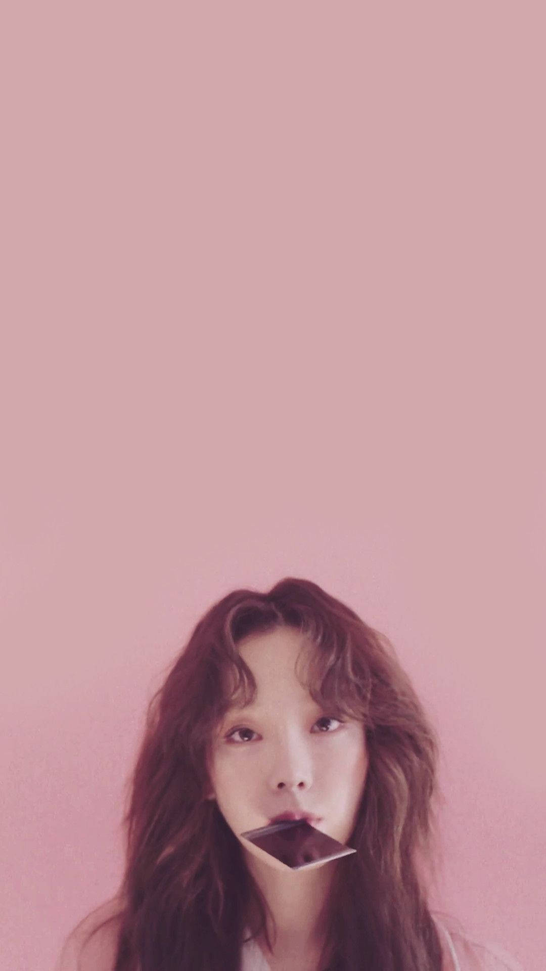 태연 桌布 壁紙 — 一位年輕女性，波浪形棕色頭髮，嘴裡含著一條黑巧克力棒，背景為純粉紅色。