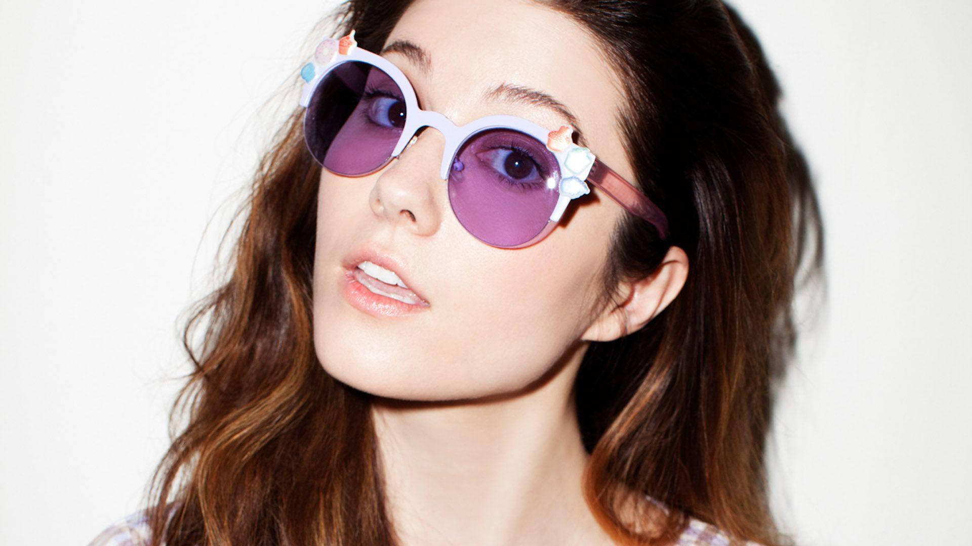 Sunglasses 桌布 壁紙 — 一位年輕女性，波浪形棕色頭髮，戴著帶有花卉裝飾的超大紫色太陽眼鏡，背景為純白色。