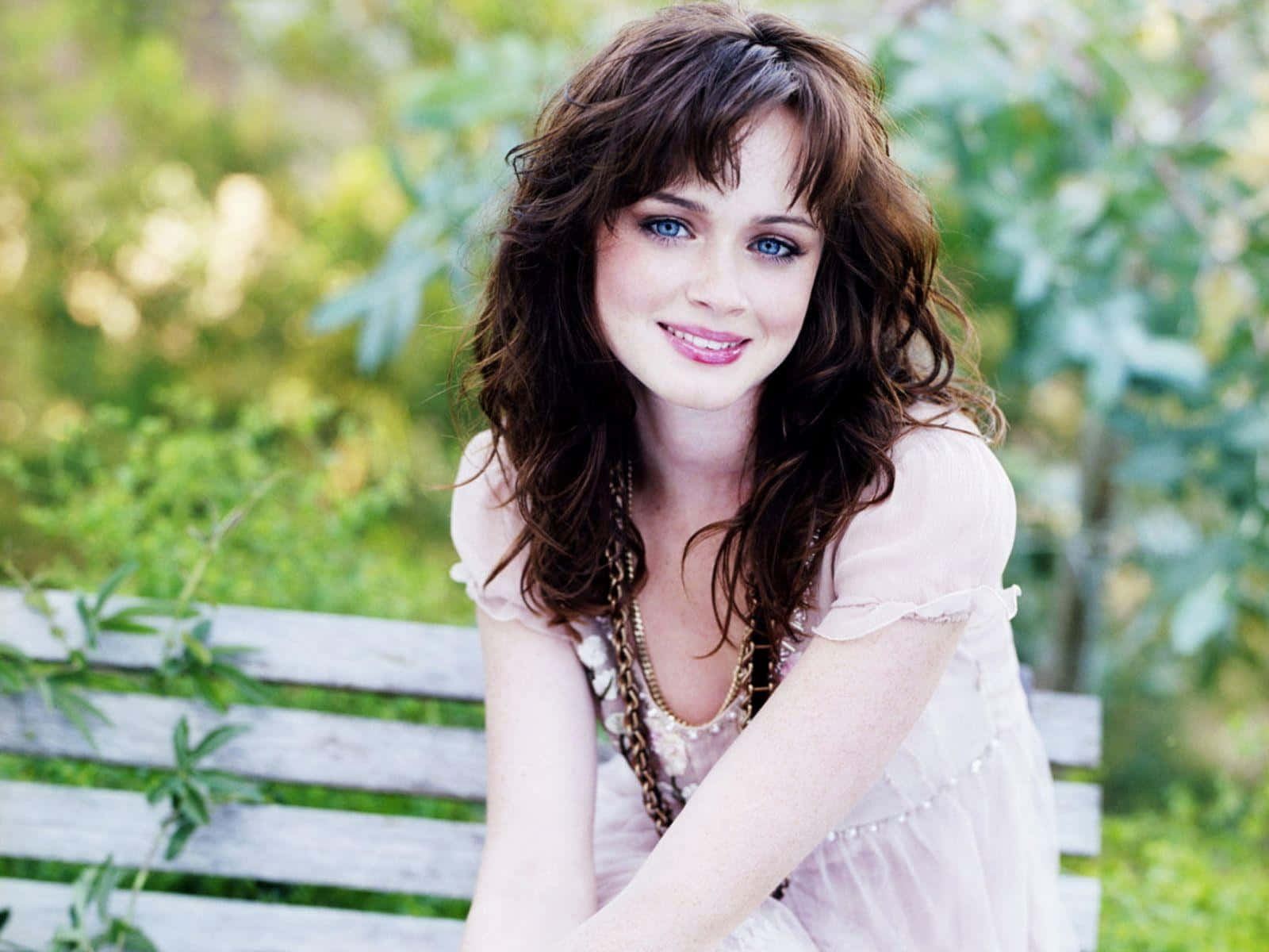 Alexis Bledel 桌布 壁紙 — 一位年輕女性，波浪形棕色頭髮，明亮的藍色眼睛，坐在木長椅上，周圍是茂密的綠色植物，溫暖地微笑。