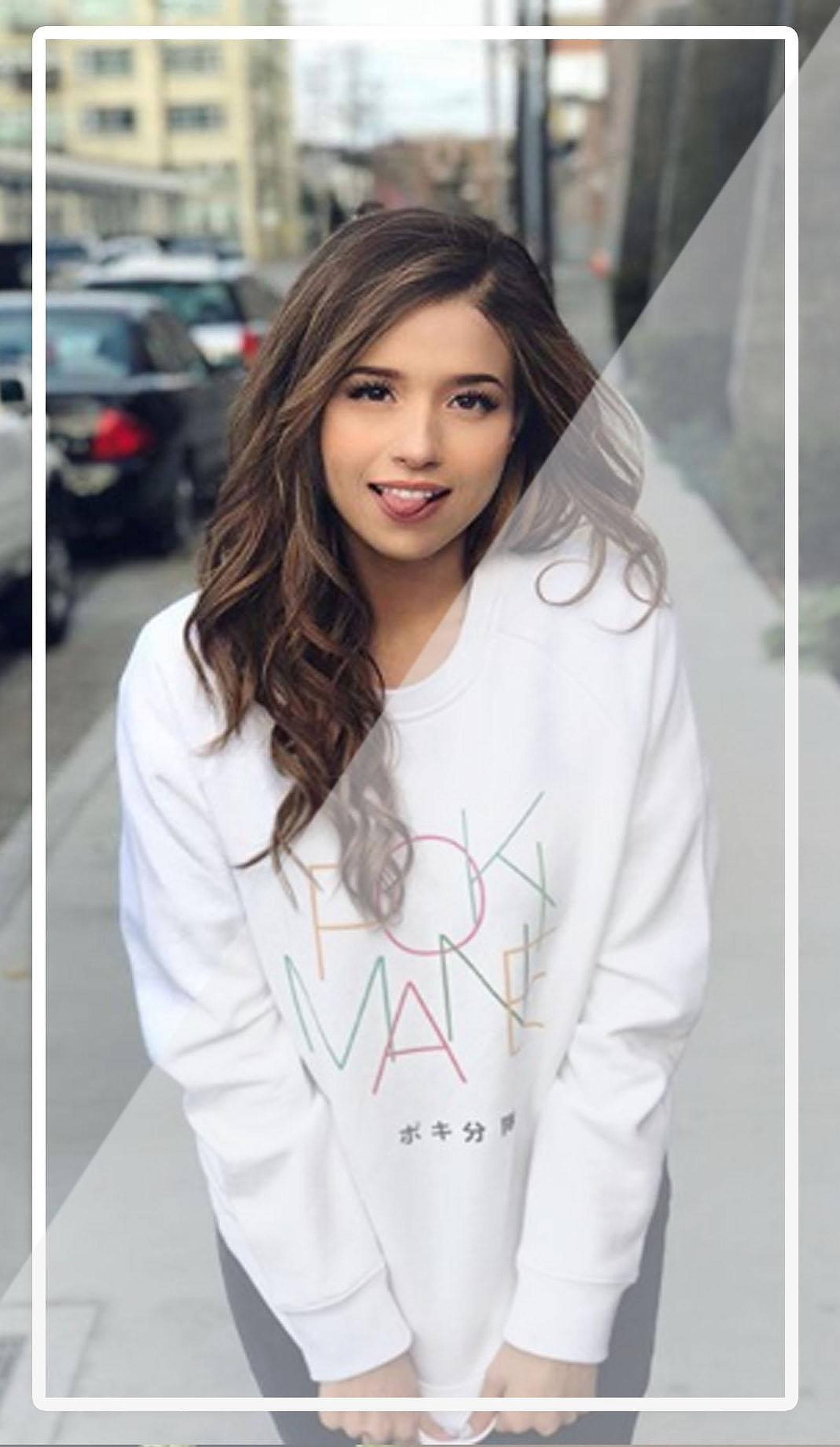 Pokimane 桌布 壁紙 — 一位年輕女性，波浪形棕色頭髮，穿著帶有彩色文字的白色運動衫，在城市人行道上微笑。