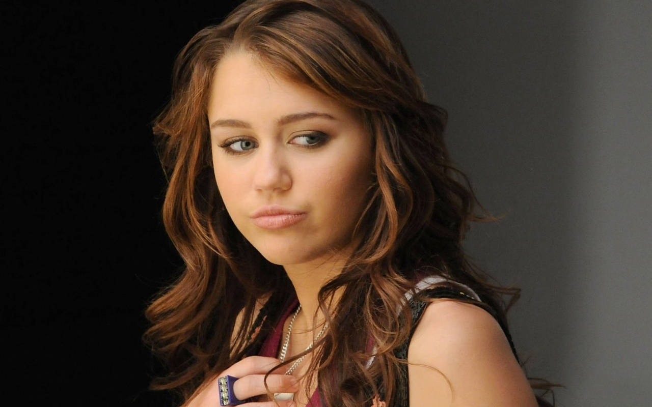 miley 桌布 壁紙 — 一位年輕女性，波浪形棕色頭髮，表情嚴肅，在深色背景下直視鏡頭。