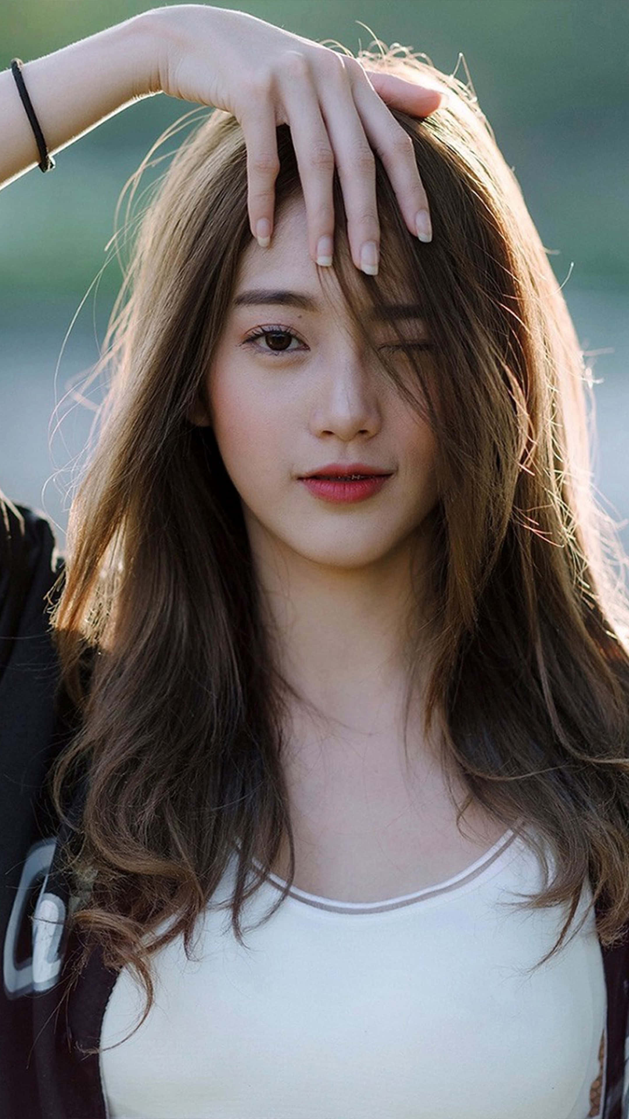 Cute Girl 桌布 壁紙 — 一位年輕女性，波浪棕色頭髮，在柔和自然光中輕觸額頭，同時向鏡頭眨眼。