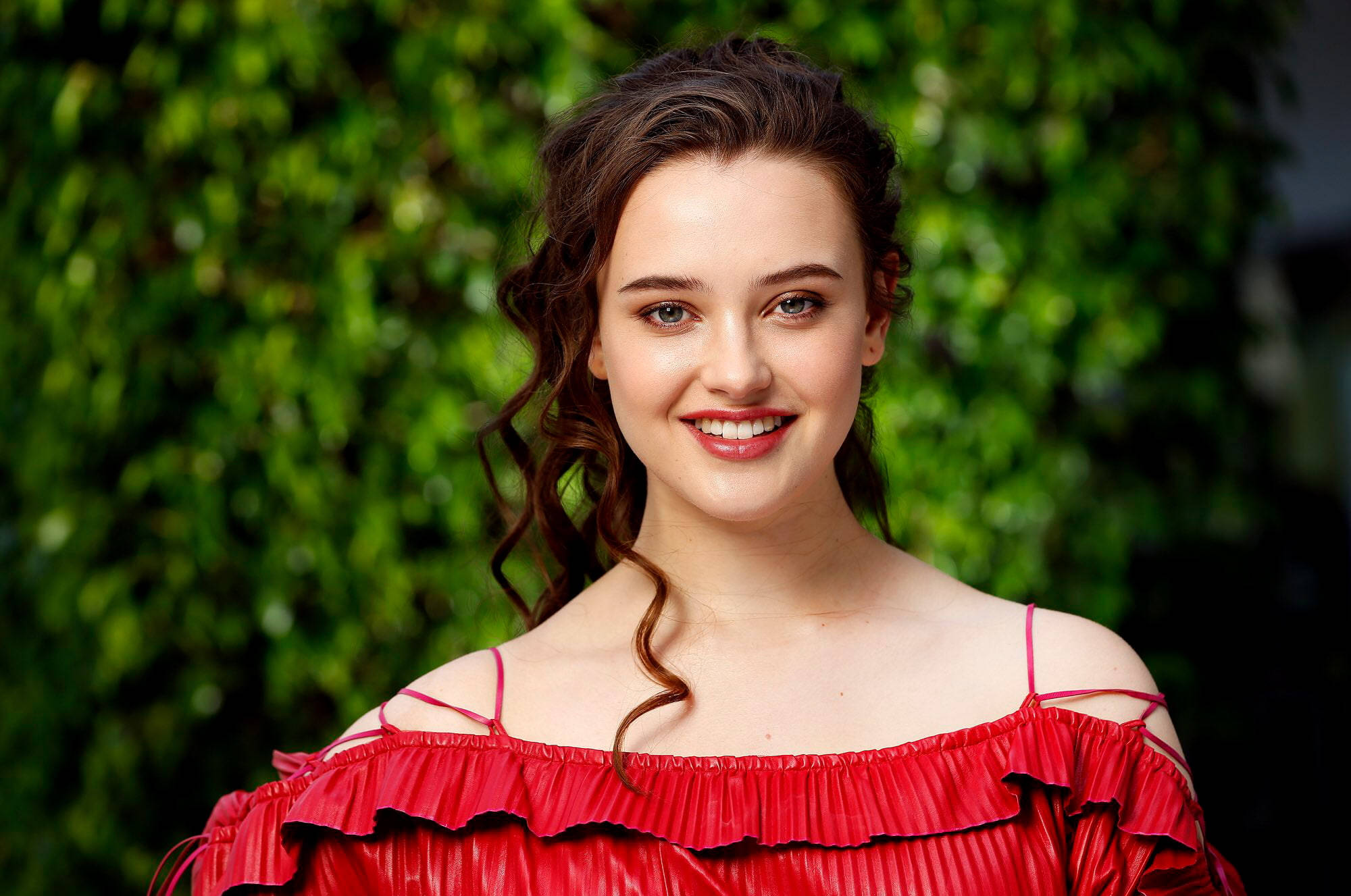 Katherine Langford 桌布 壁紙 — 一位年輕女性，波浪棕色頭髮，穿著鮮紅色露肩連衣裙，在茂密綠色樹葉背景下溫暖微笑。
