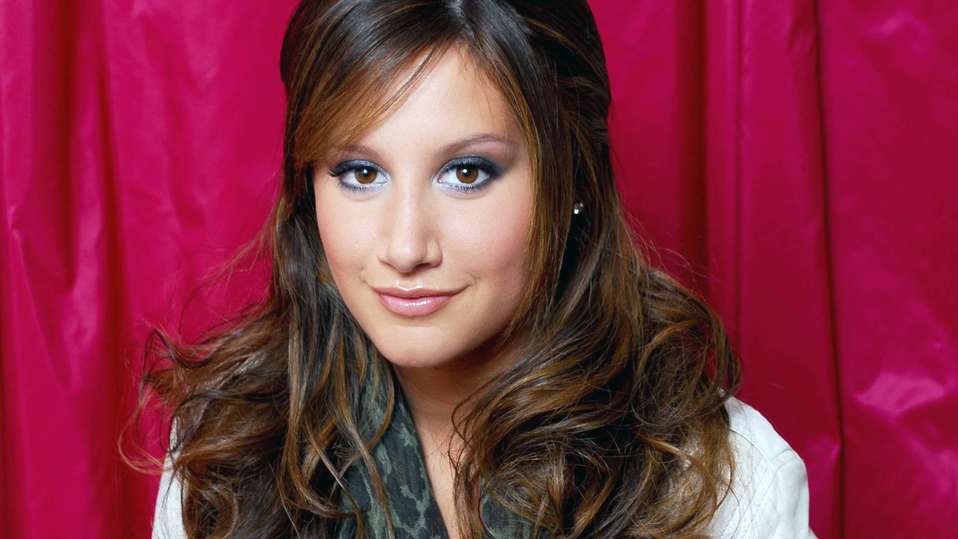Ashley Tisdale 桌布 壁紙 — 一位年輕女性，波浪棕色頭髮和煙燻眼妝，在鮮豔的粉紅色窗簾背景下溫柔微笑。