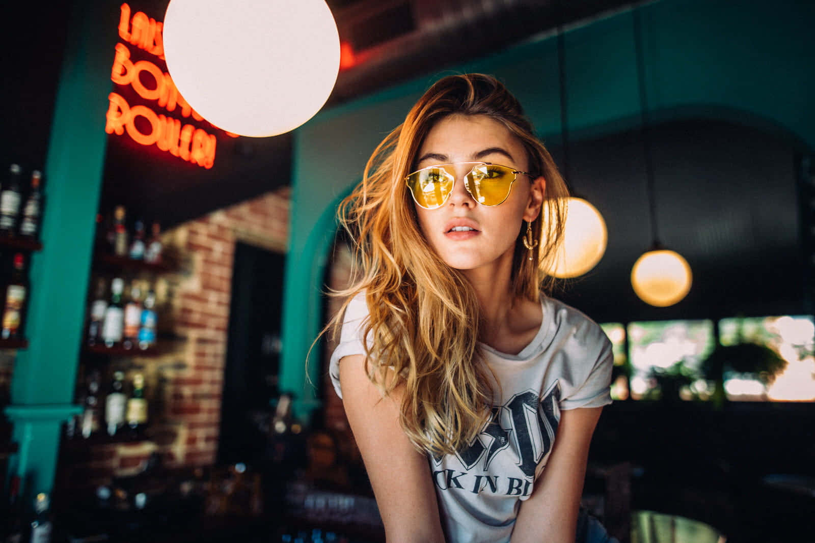 Sunglasses 桌布 壁紙 — 一位年輕女性，波浪金髮，戴著黃色太陽眼鏡和圖案T恤，坐在光線昏暗的酒吧裡，周圍有霓虹燈招牌和吊燈。