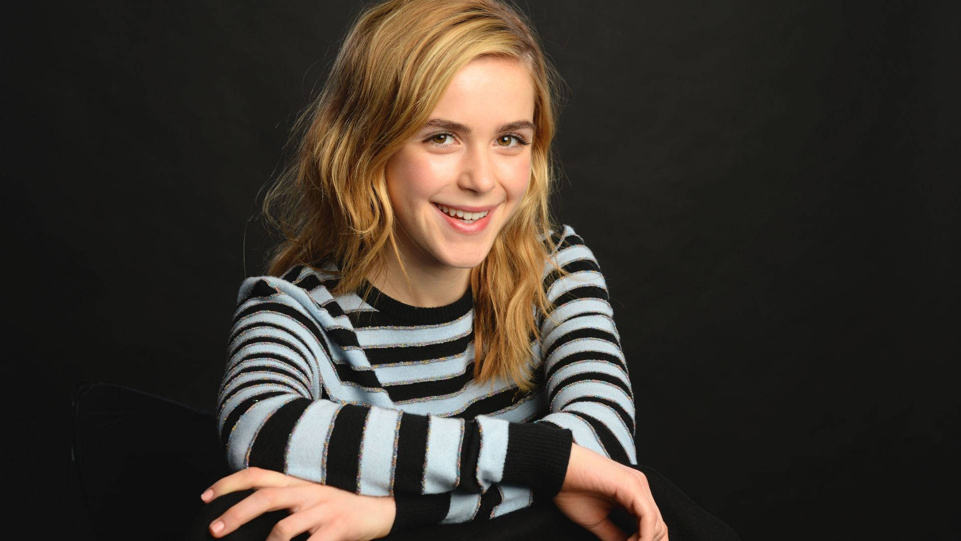 Kiernan Shipka 桌布 壁紙 — 一位年輕女性，波浪金髮，穿著黑色和淺藍色條紋毛衣，在深色背景下溫暖微笑。