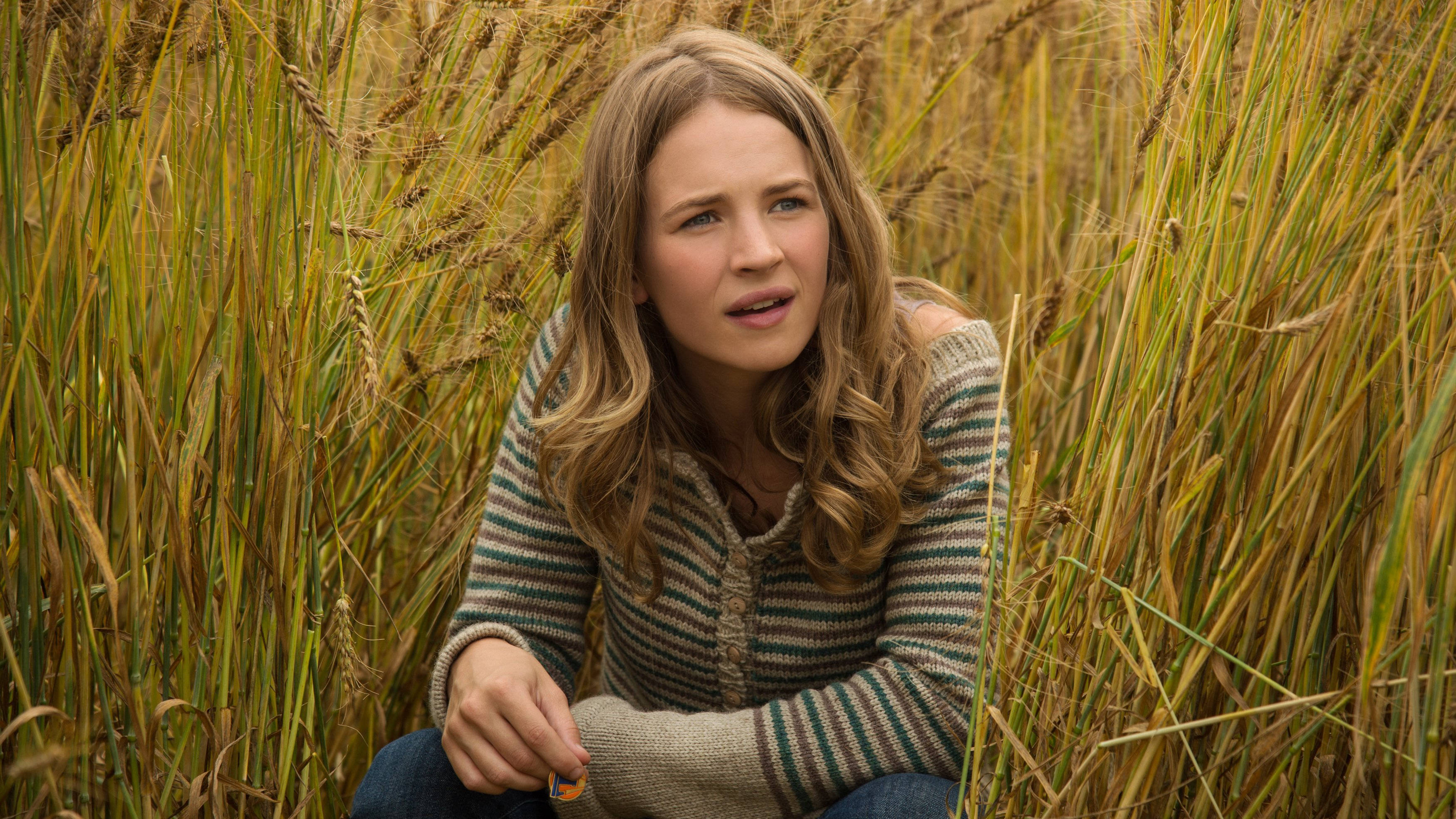 Tomorrowland Movie 桌布 壁紙 — 一位年輕女性，波浪金髮，身穿條紋毛衣，坐在金色麥田裡，目光望向鏡頭外，神情若有所思。