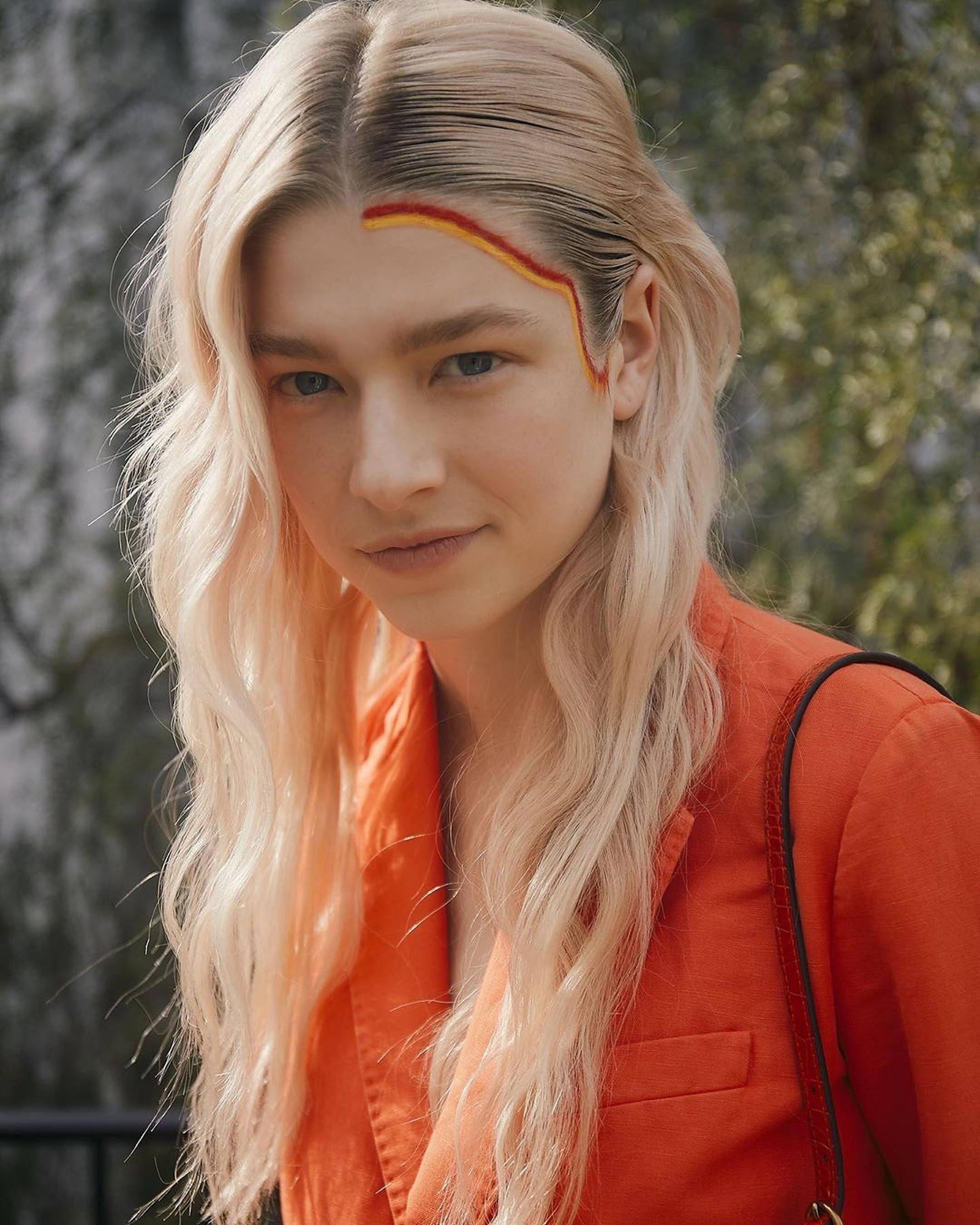 Hunter Schafer 桌布 壁紙 — 一位年輕女性，波浪金髮，額頭有紅黃條紋，戶外穿著橙色外套。