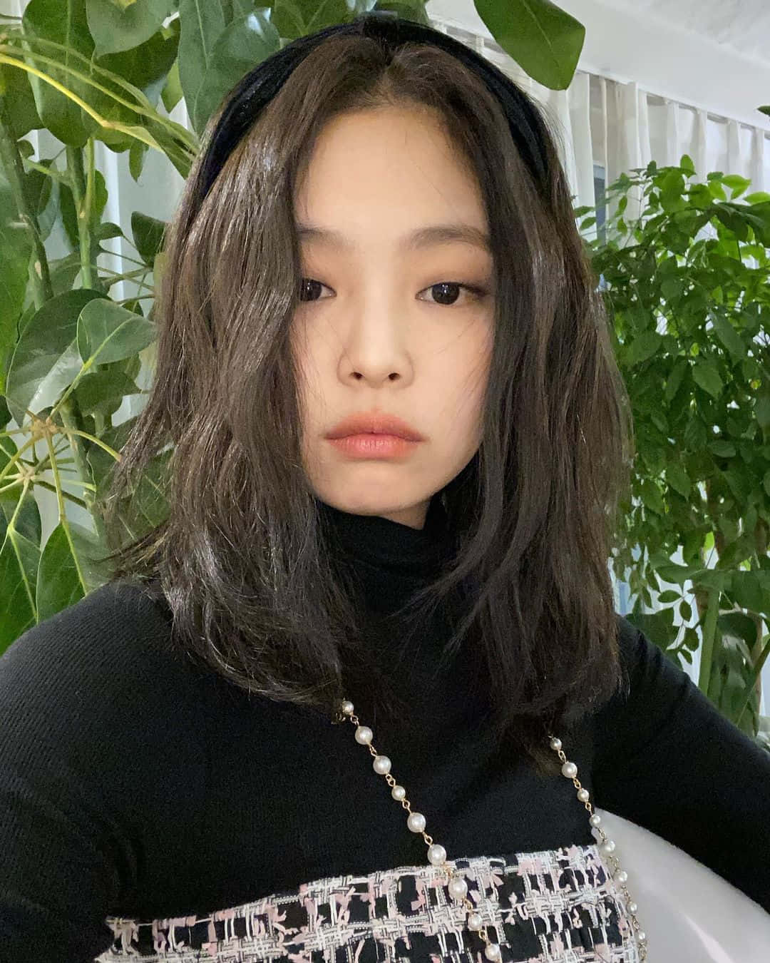 BLACKPINK 桌布 壁紙 — 一位年輕女性，深色波浪長髮搭配黑色髮帶，身穿黑色高領衫，手持珍珠裝飾手袋，在茂密綠葉前溫柔凝視。
