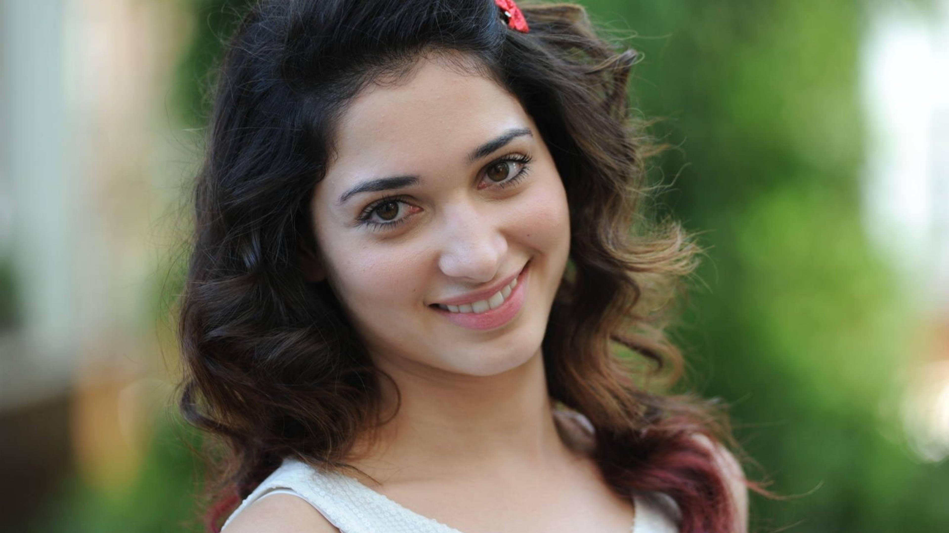 tamannaah bhatia 桌布 壁紙 — 一位年輕女性，深色波浪頭髮，紅色髮夾，在柔和模糊的綠色背景前溫暖微笑。