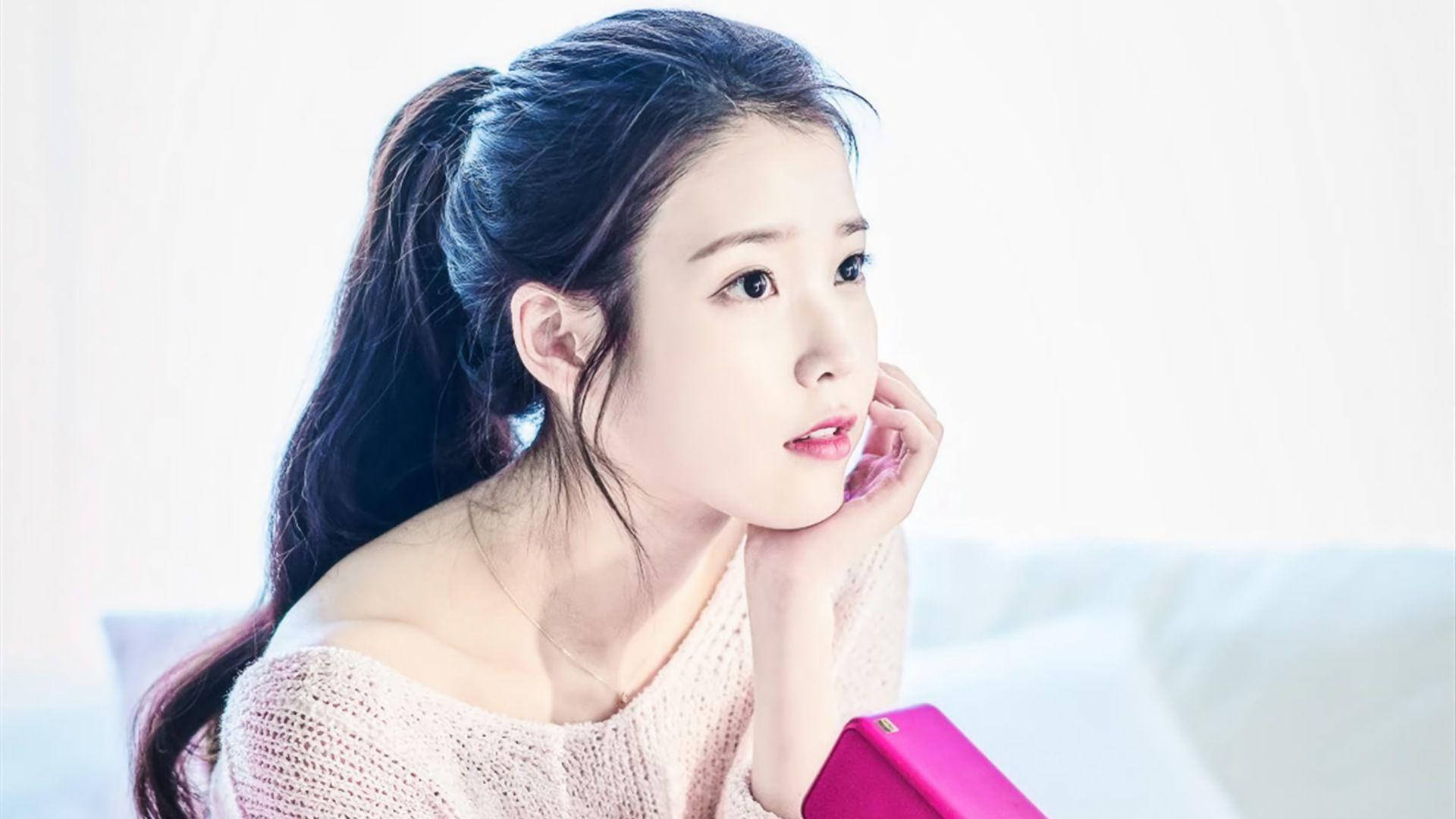 Kpop Aesthetic 桌布 壁紙 — 一位年輕女性，深色頭髮紮成馬尾辮，托腮沉思，身穿柔軟的粉紅色針織毛衣。