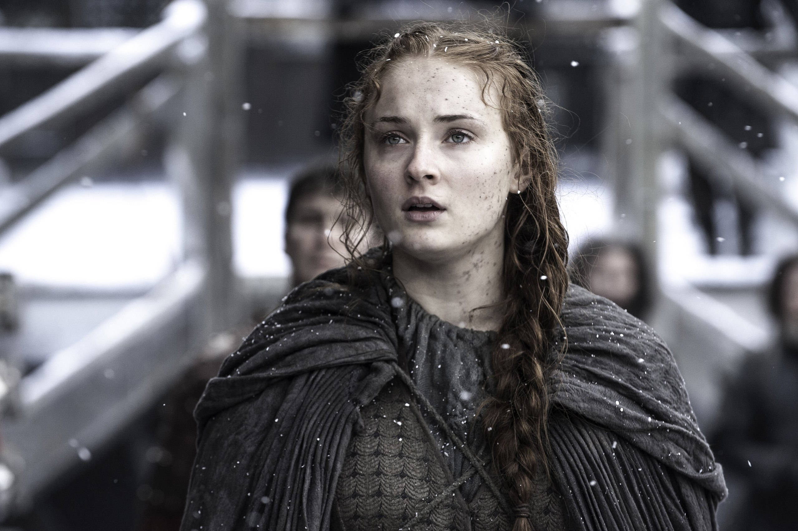 Sansa Stark 桌布 壁紙 — 一位年輕女性，濕辮髮，深色斗篷上有雪花，站在積雪的金属結構中，表情驚訝。