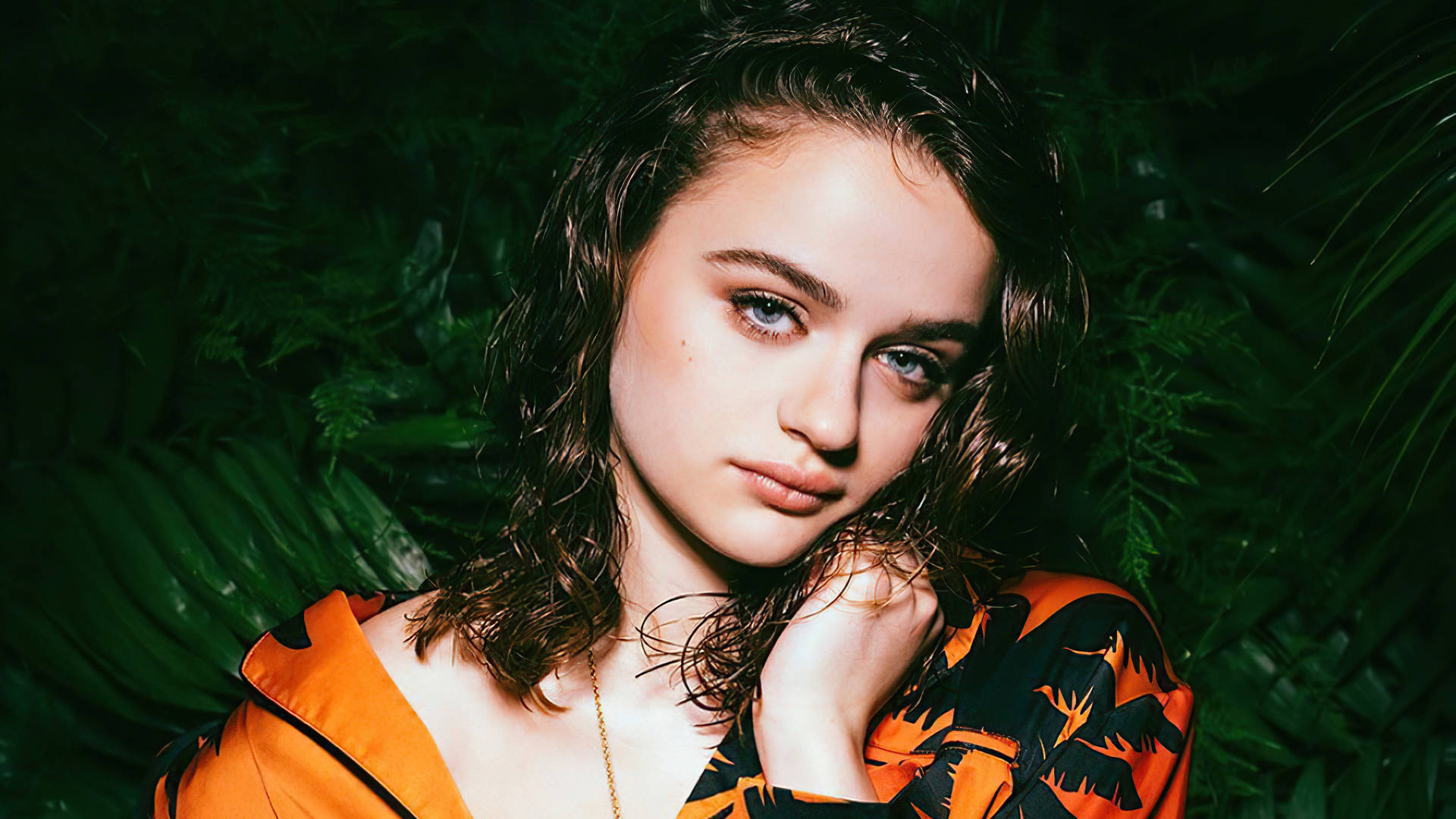 Joey King 桌布 壁紙 — 一位年輕女性，濕髮，引人注目的藍色眼睛，穿著橙色熱帶印花上衣，背景是茂密的綠色樹葉，溫柔地凝視著。