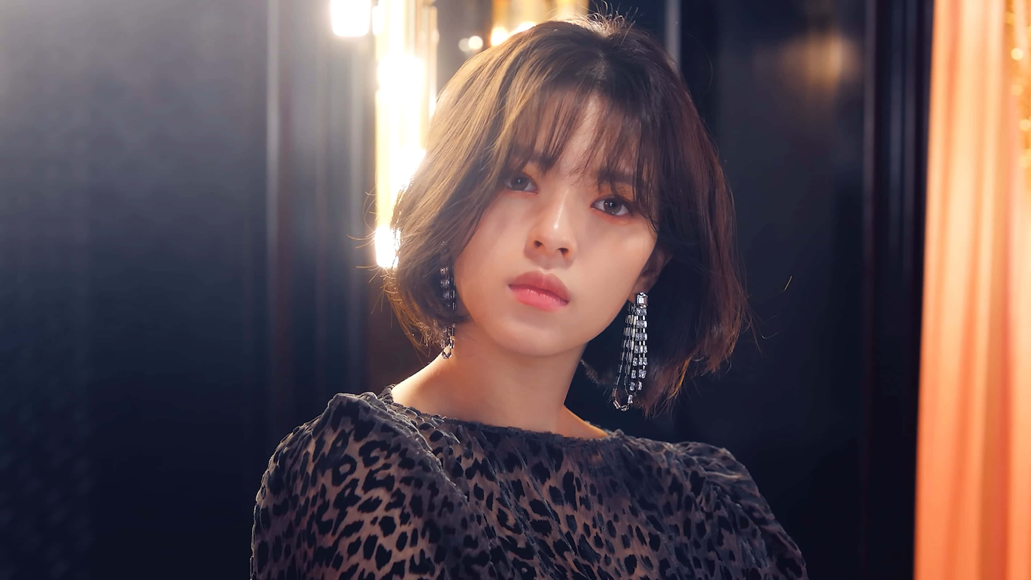 twice 桌布 壁紙 — 一位年輕女性，留著短棕髮，戴著大膽的耳環，在昏暗的房間裡，身後有溫暖發光的燈光，直視鏡頭。