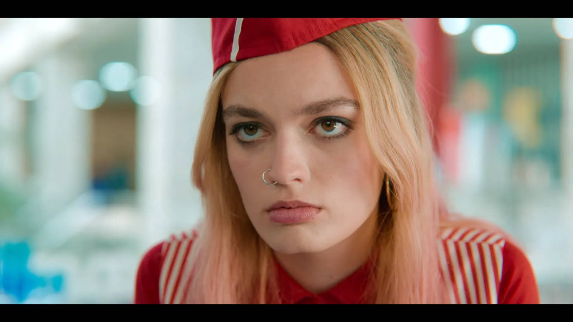 maeve wiley 桌布 壁紙 — 一位年輕女性，粉紅色頭髮，身穿紅色制服，在光線明亮的室內環境中專注地凝視鏡頭。