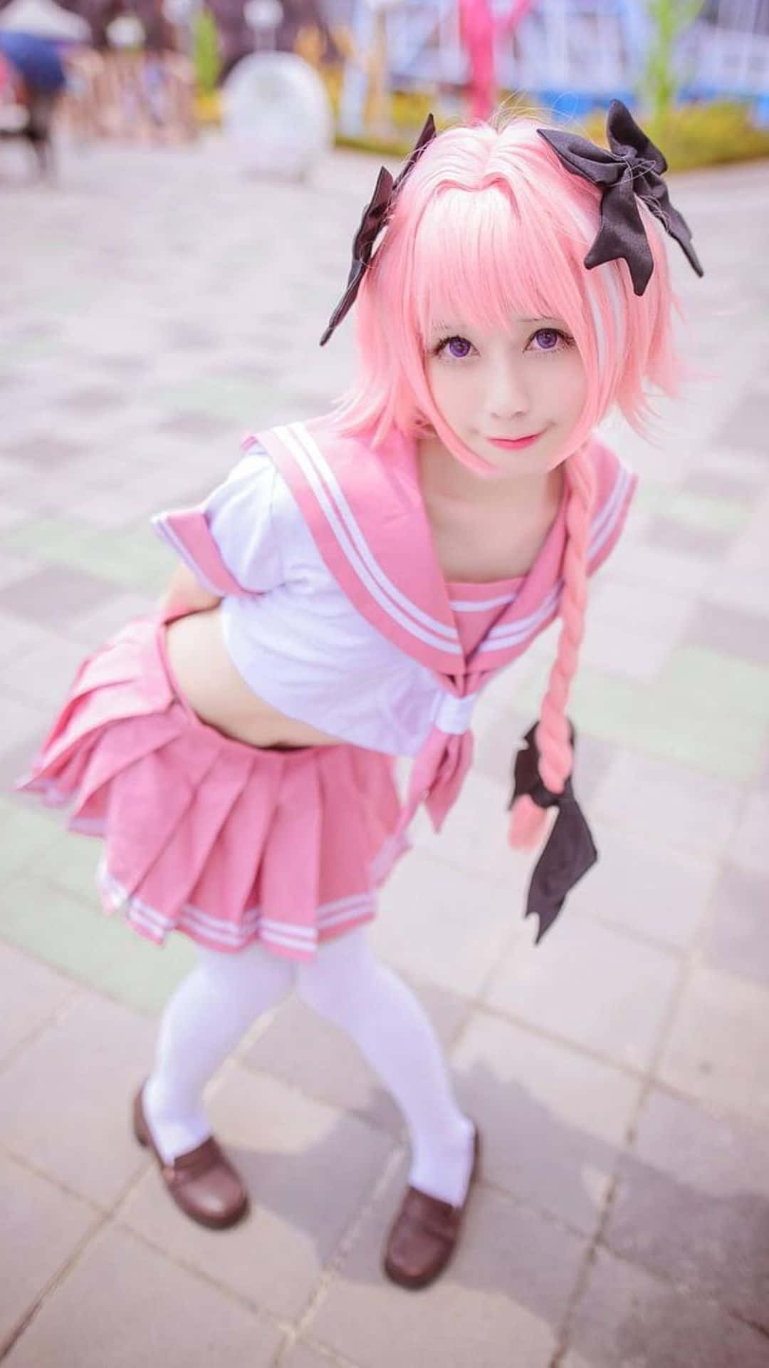 Cosplay Anime Kawaii 桌布 壁紙 — 一位年輕女性，粉紅色頭髮和黑色蝴蝶結，在瓷磚庭院中穿著粉紅色水手服擺姿勢。