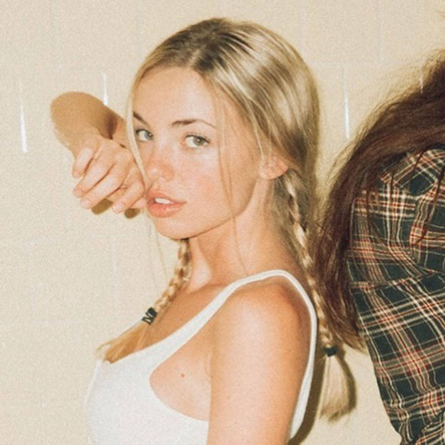 Emma Kotos 桌布 壁紙 — 一位年輕女性，金色辮子，身穿白色背心，在素牆前將手靠近臉龐擺姿勢。