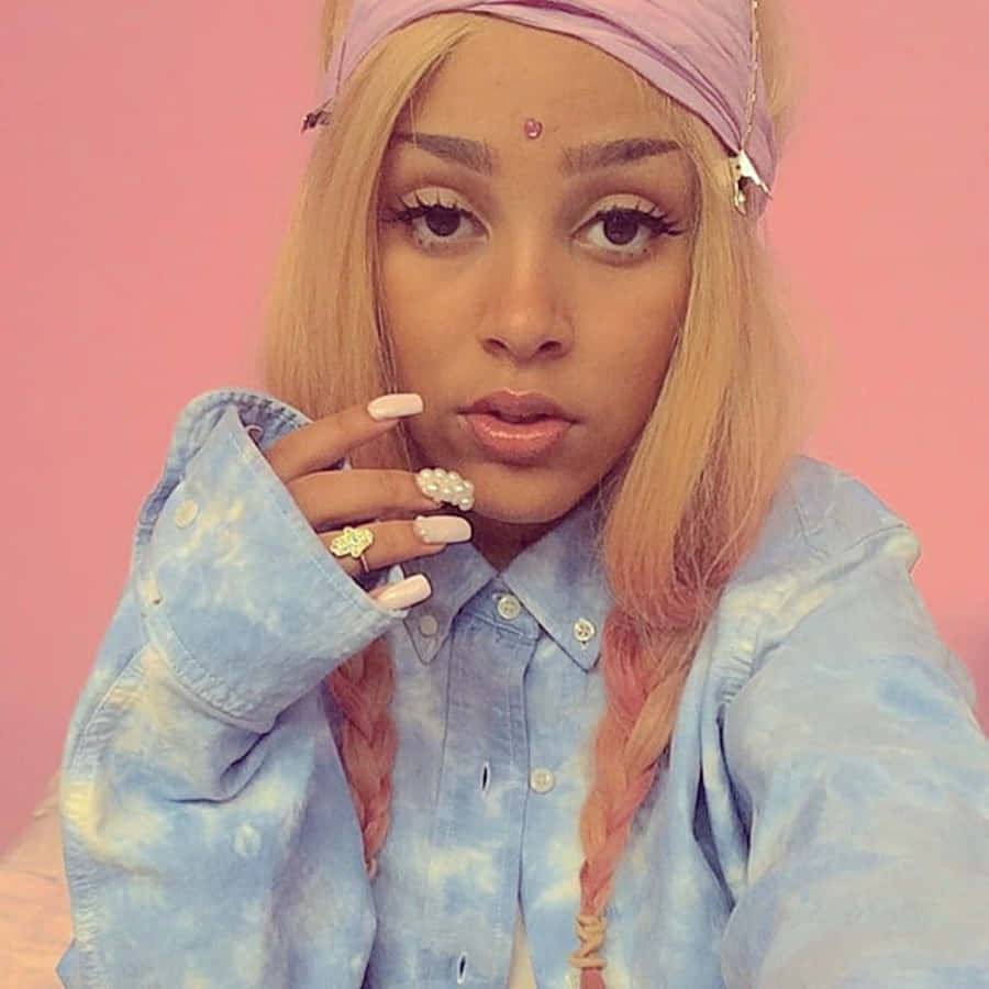 Doja Cat 桌布 壁紙 — 一位年輕女性，金色辮子配粉紅色頭帶，身穿紮染牛仔襯衫，在純粉紅色背景前自信擺姿勢。