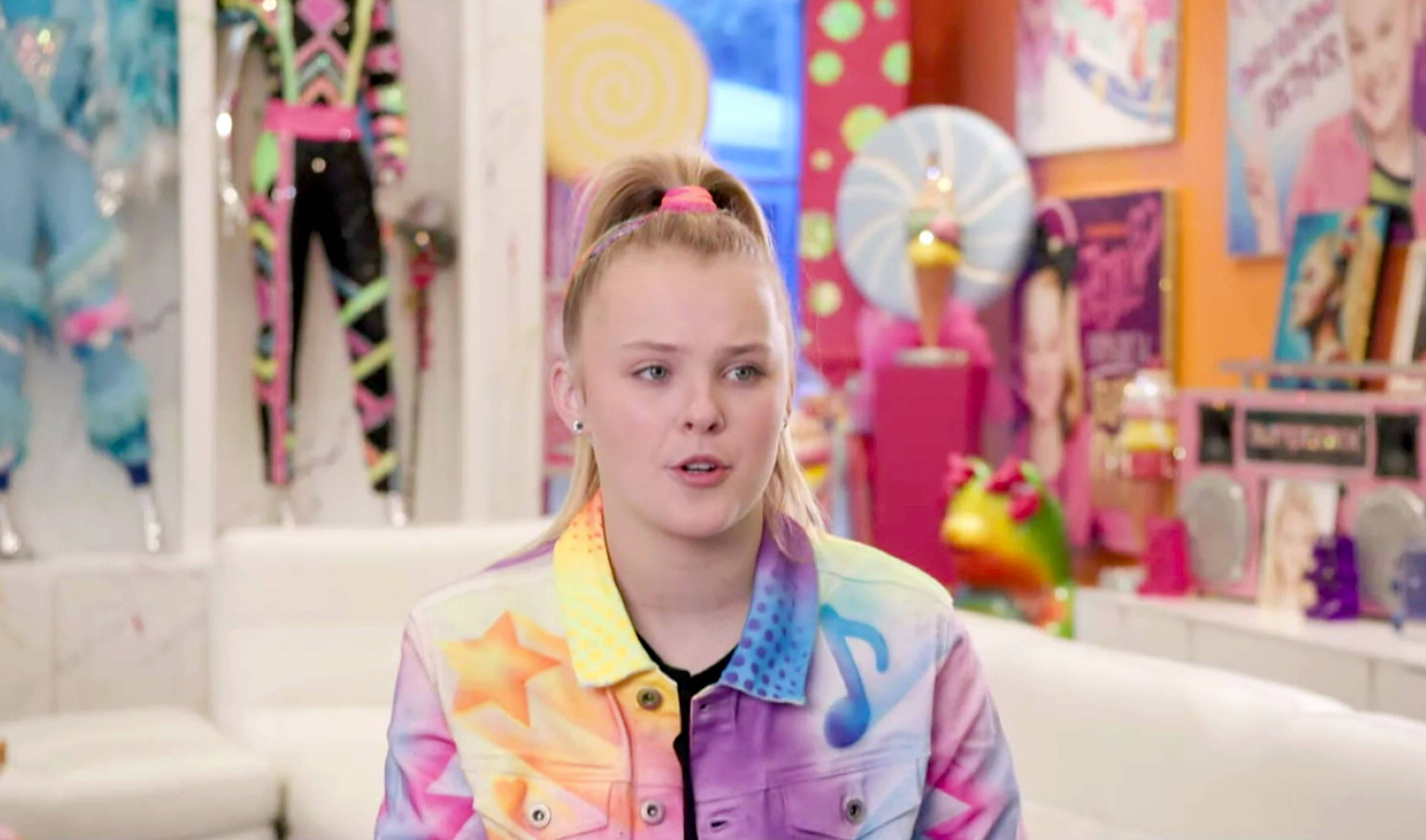 JoJo Siwa 桌布 壁紙 — 一位年輕女性，金色頭髮紮成雙馬尾，身穿帶有星星和音符的彩色外套，身處充滿活力的糖果主題房間。