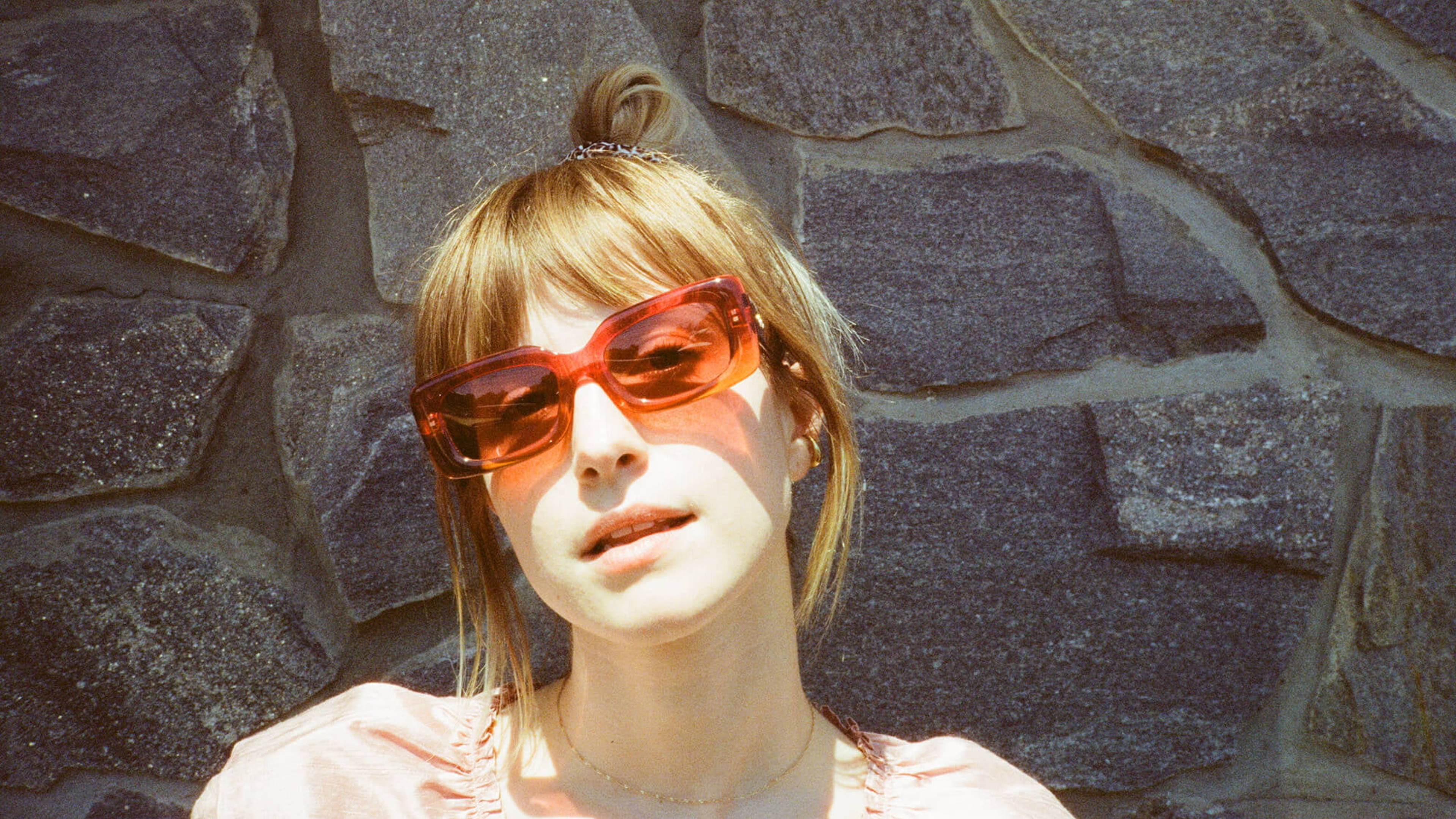 Hayley Williams 桌布 壁紙 — 一位年輕女性，金髮盤成髮髻，戴著大膽的紅色太陽眼鏡，在明亮的陽光下以有紋理的石牆為背景。