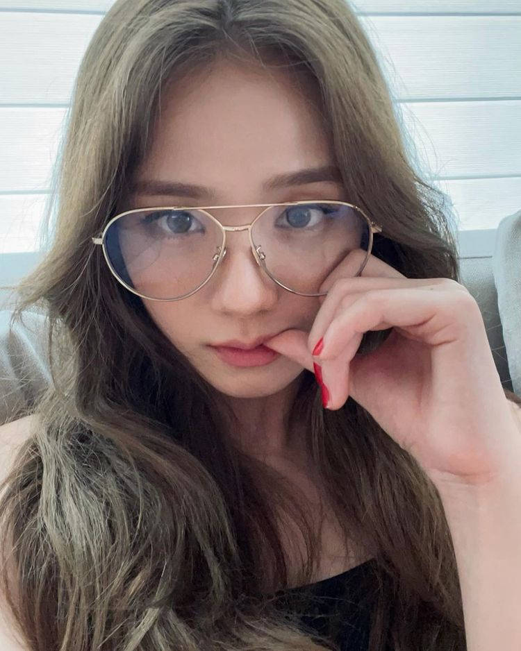 Jisoo Cute 桌布 壁紙 — 一位年輕女性，長波浪頭髮，戴金框眼鏡，手靠近臉龐，以明亮的窗戶為背景擺姿勢。