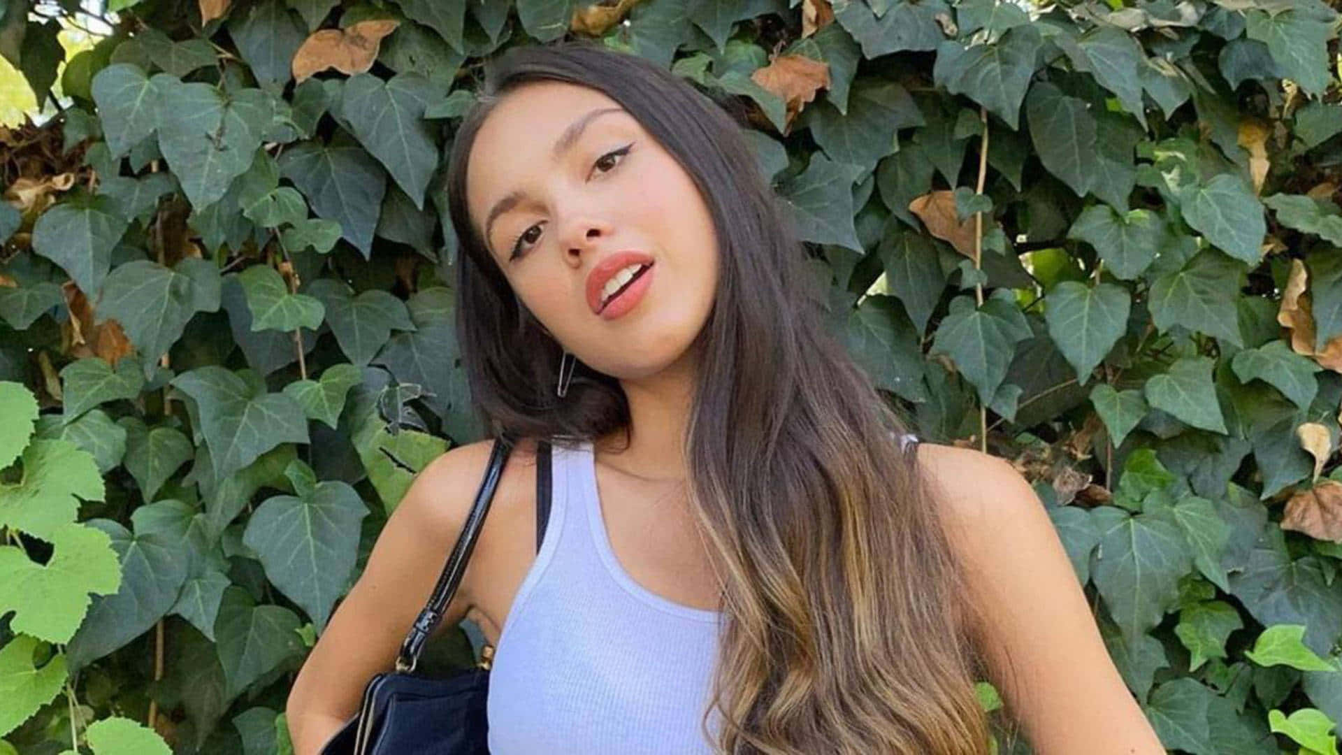 奧莉維亞·羅德里戈 桌布 壁紙 — 一位年輕女性，長著深色長髮，穿著淺藍色背心，自信地在茂密的綠色常春藤牆前擺姿勢。