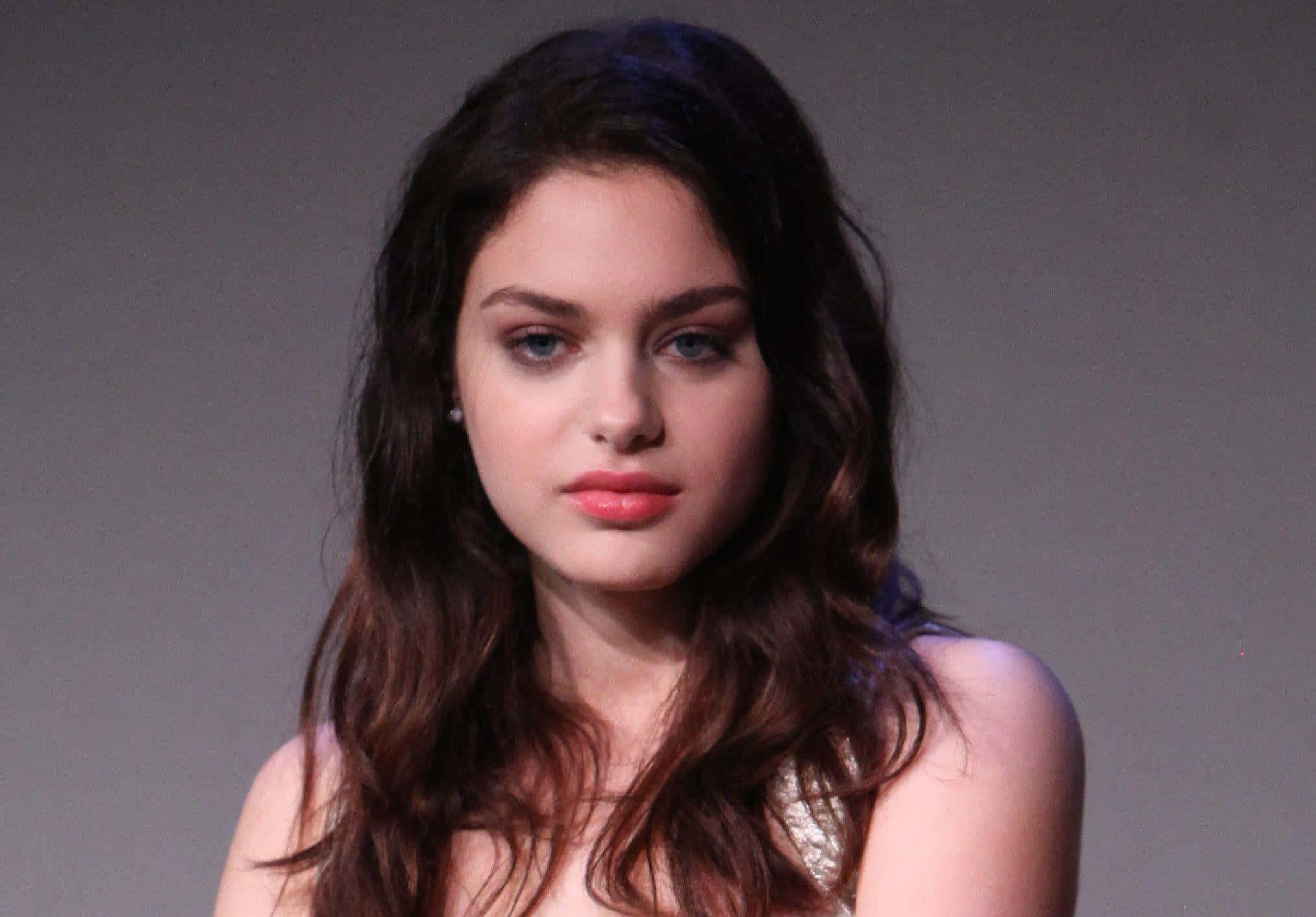 Odeya Rush 桌布 壁紙 — 一位年輕女性，長著長長的波浪形棕色頭髮和引人注目的藍色眼睛，在純灰色背景前直視鏡頭。