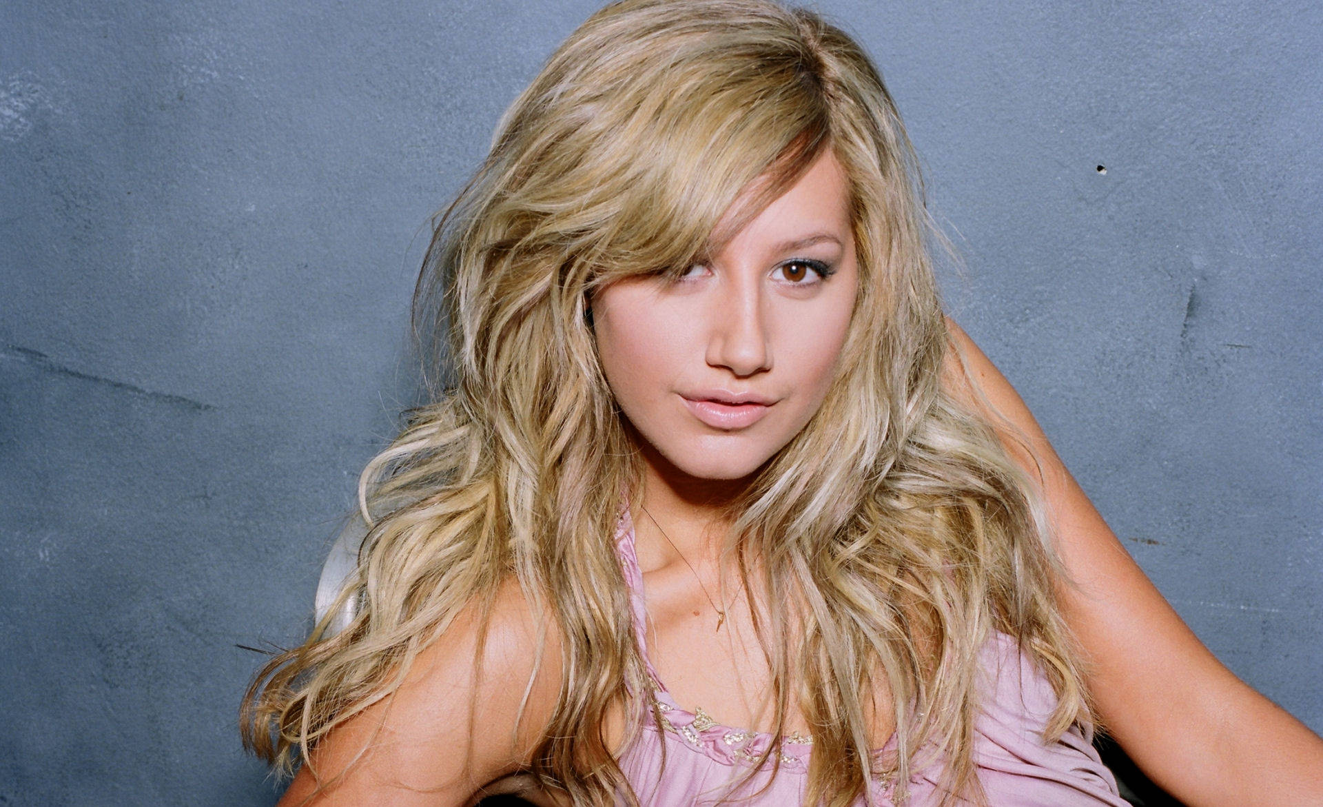 Ashley Tisdale 桌布 壁紙 — 一位年輕女性，長著長長的波浪金髮，身穿粉紅色上衣，在紋理灰色牆壁前擺姿勢。