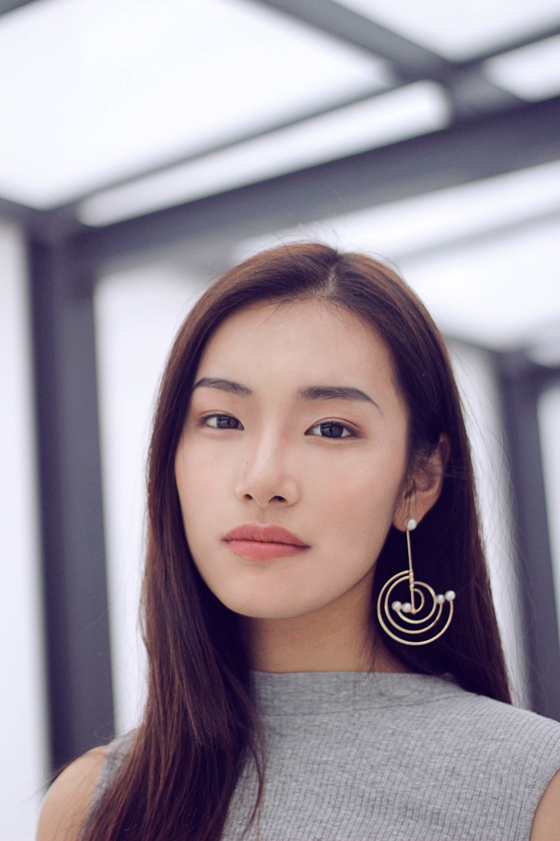 lovely girl 桌布 壁紙 — 一位年輕女性，長長棕色頭髮，戴著時尚螺旋耳環，在現代玻璃和鋼結構下平靜地凝視鏡頭。