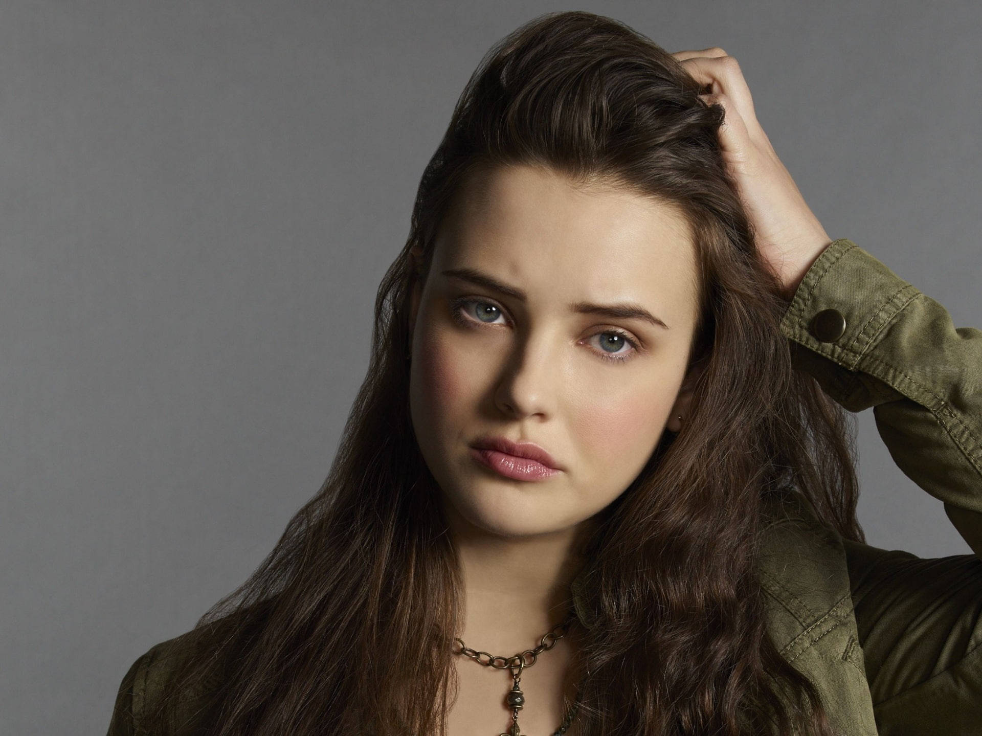 Katherine Langford 桌布 壁紙 — 一位年輕女性，長長棕色頭髮和引人注目的藍色眼睛，直視鏡頭，一手觸摸頭髮，身穿橄欖綠外套，背景為純灰色。