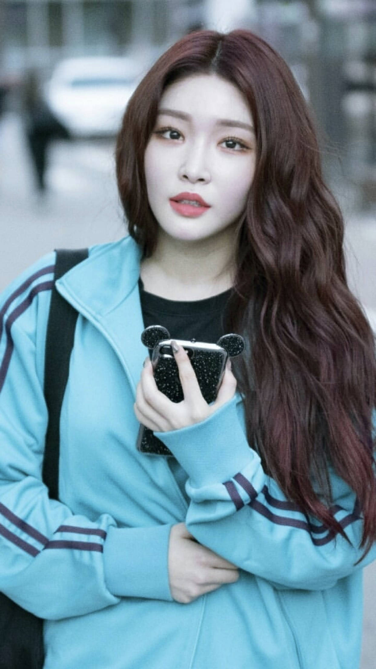 Chungha 桌布 壁紙 — 一位年輕女性，長長波浪赤褐色頭髮，穿著淺藍色運動外套，手持閃亮手機殼，站在戶外。
