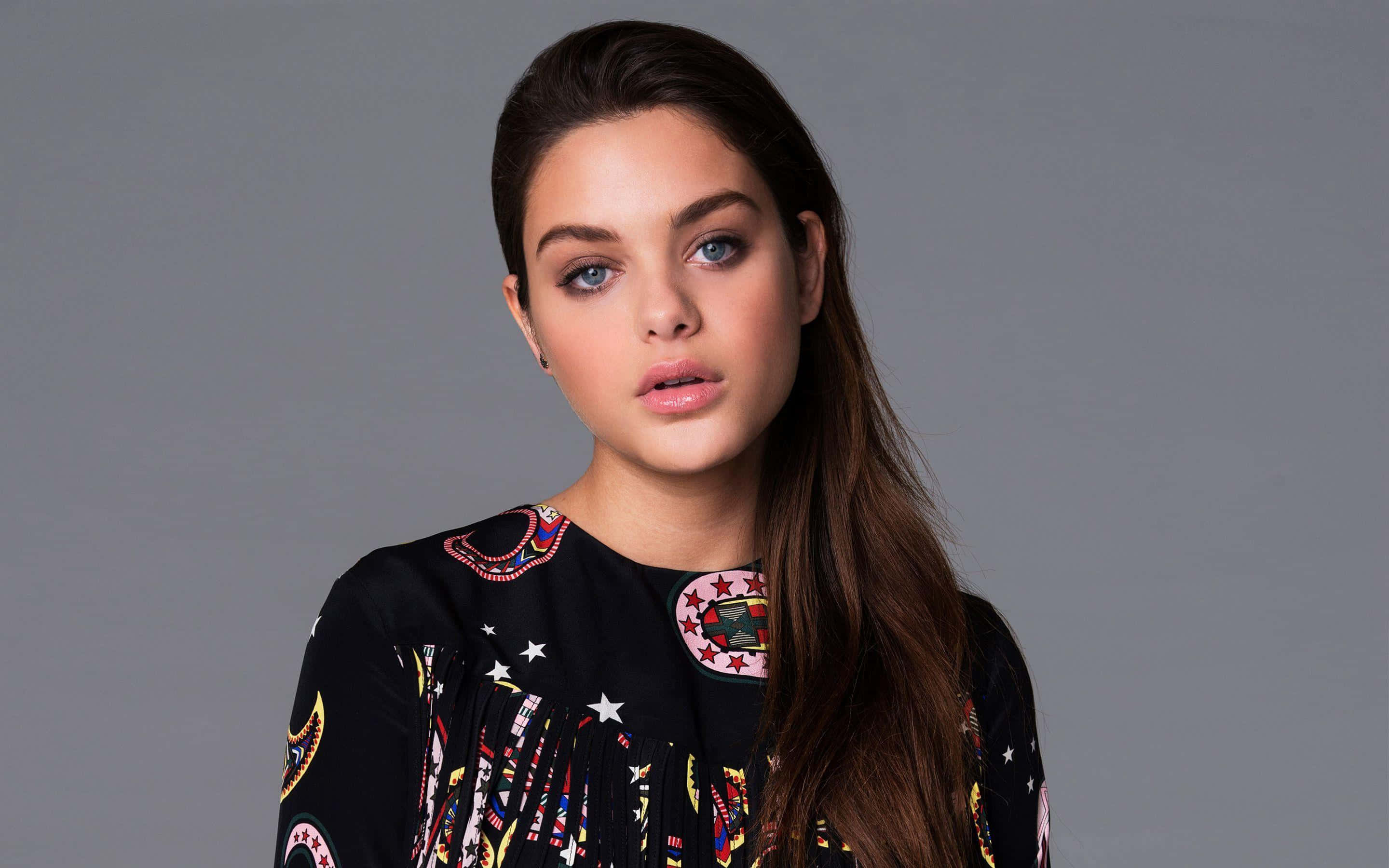 Odeya Rush 桌布 壁紙 — 一位年輕女性，長長的棕色頭髮和引人注目的藍色眼睛，直視鏡頭，穿著黑色圖案上衣，背景是純灰色。