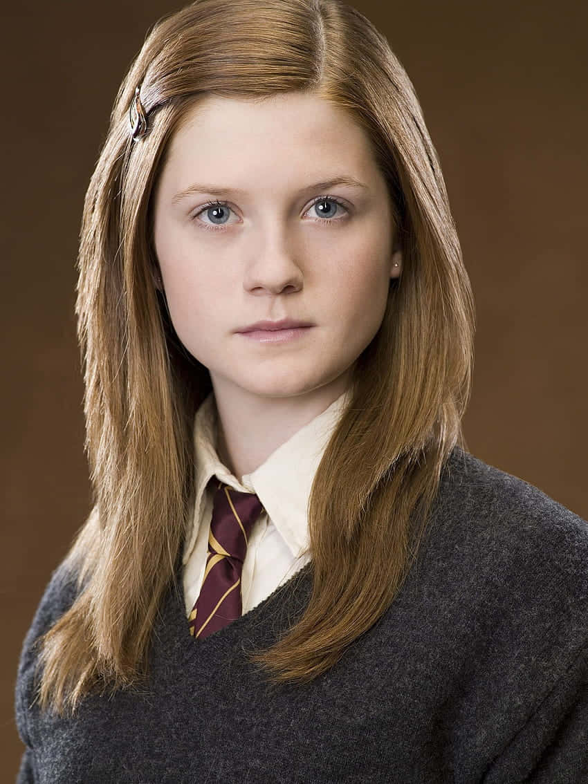 Ginny Weasley 桌布 壁紙 — 一位年輕女性，長長的棕色頭髮和藍色眼睛，穿著灰色毛衣和棗紅色領帶，背景是棕色。
