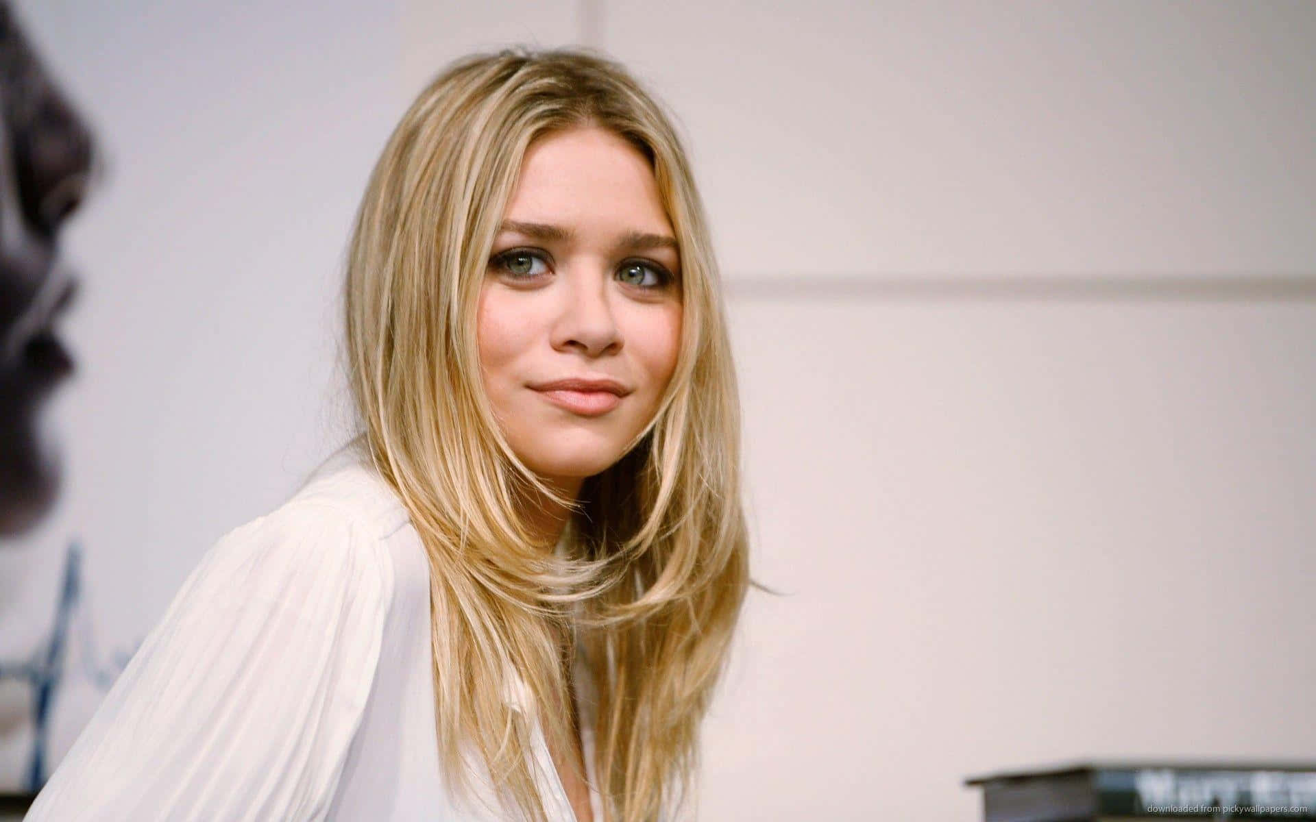 Mary Kate Olsen 桌布 壁紙 — 一位年輕女性，長長的金色頭髮，帶著溫柔的微笑，穿著白色襯衫，背景是純色淺色。