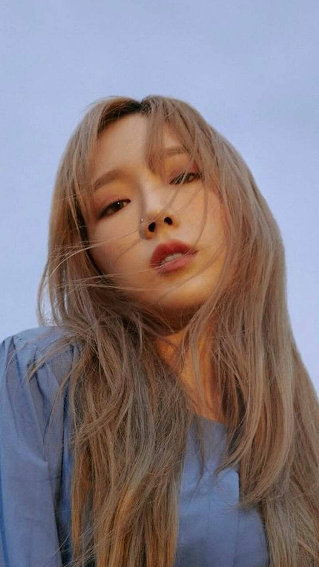 태연 桌布 壁紙 — 一位年輕女性，長長的金色頭髮和鼻環，在微風吹起她的髮絲時，溫柔地凝視著淡藍色的天空。